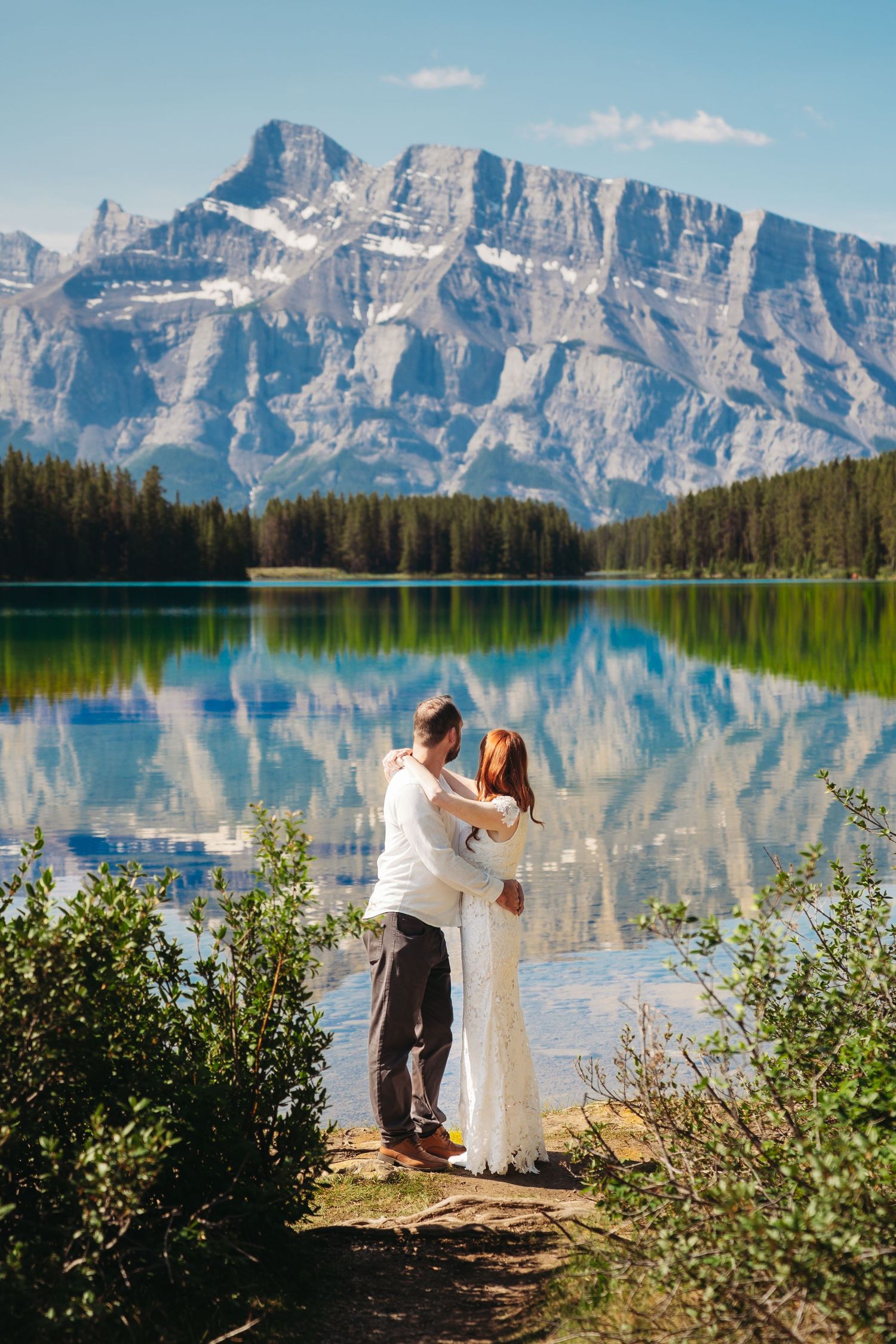 Bride and groom stand together overlooking a calm Banff lake with crystal reflections and dramatic Rocky Mountain peaks beneath a clear sky.
