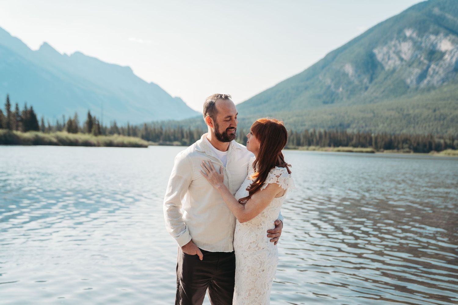 Bride and groom stand smiling by a calm mountain lake in Banff, surrounded by alpine peaks, evergreen trees, and clear blue summer skies.