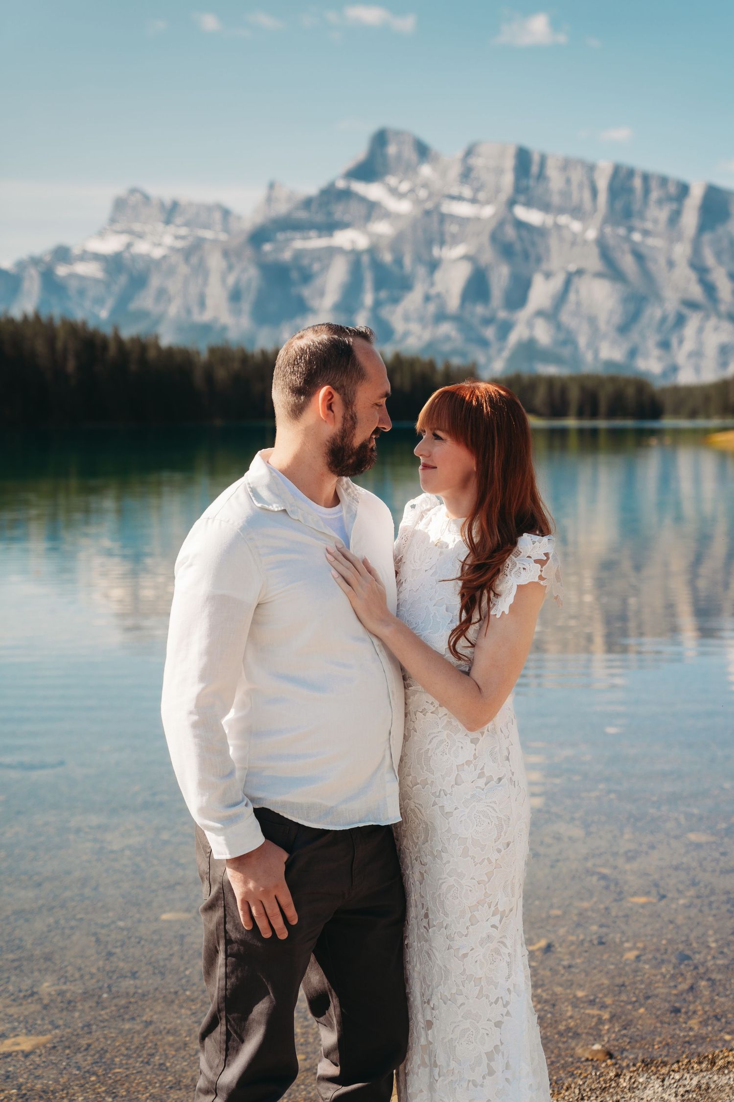 Bride and groom standing by a clear mountain lake in Banff, sharing an intimate moment with sunlit peaks and reflections behind them.