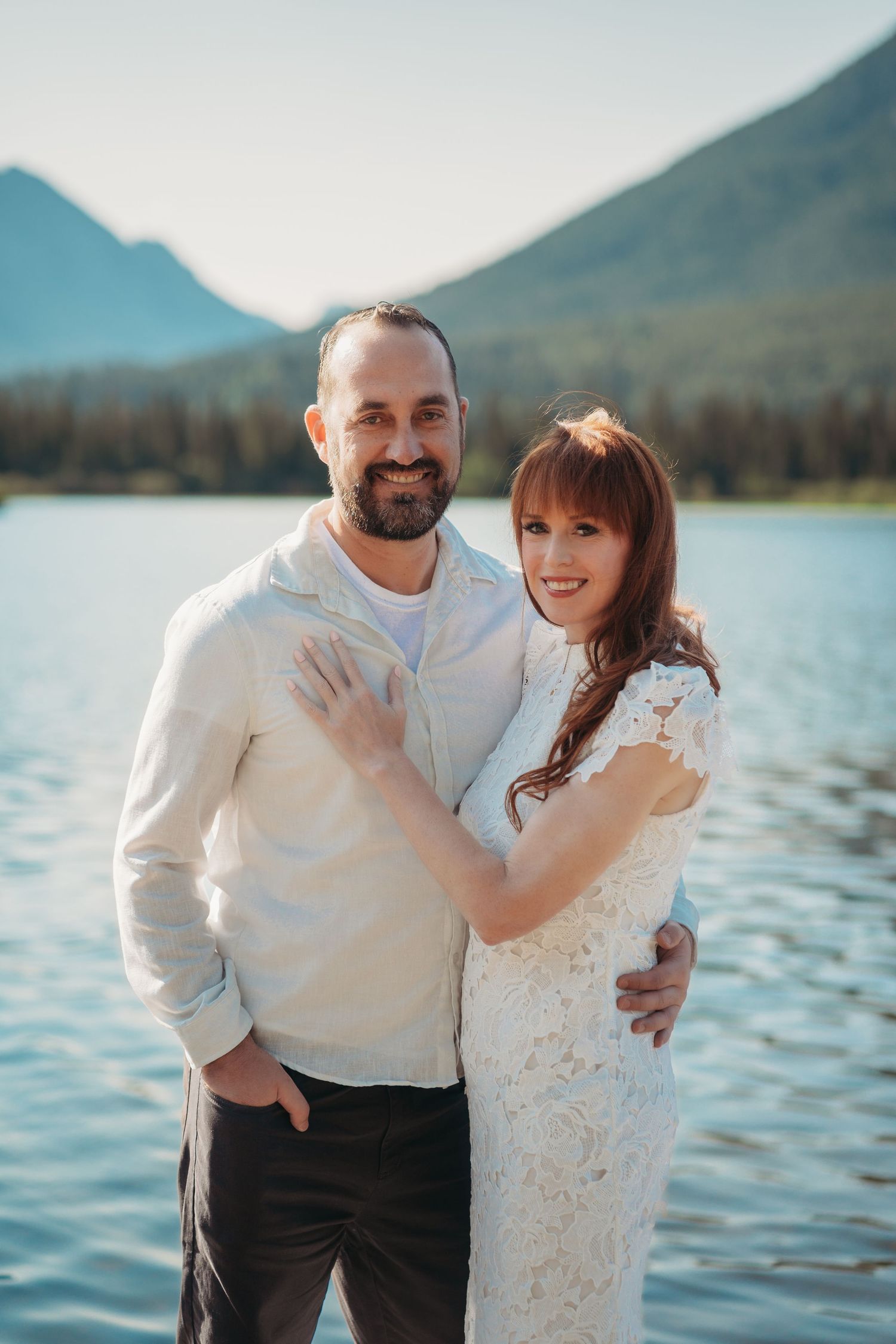 Bride and groom stand together overlooking a calm Banff lake with crystal reflections and dramatic Rocky Mountain peaks beneath a clear sky.