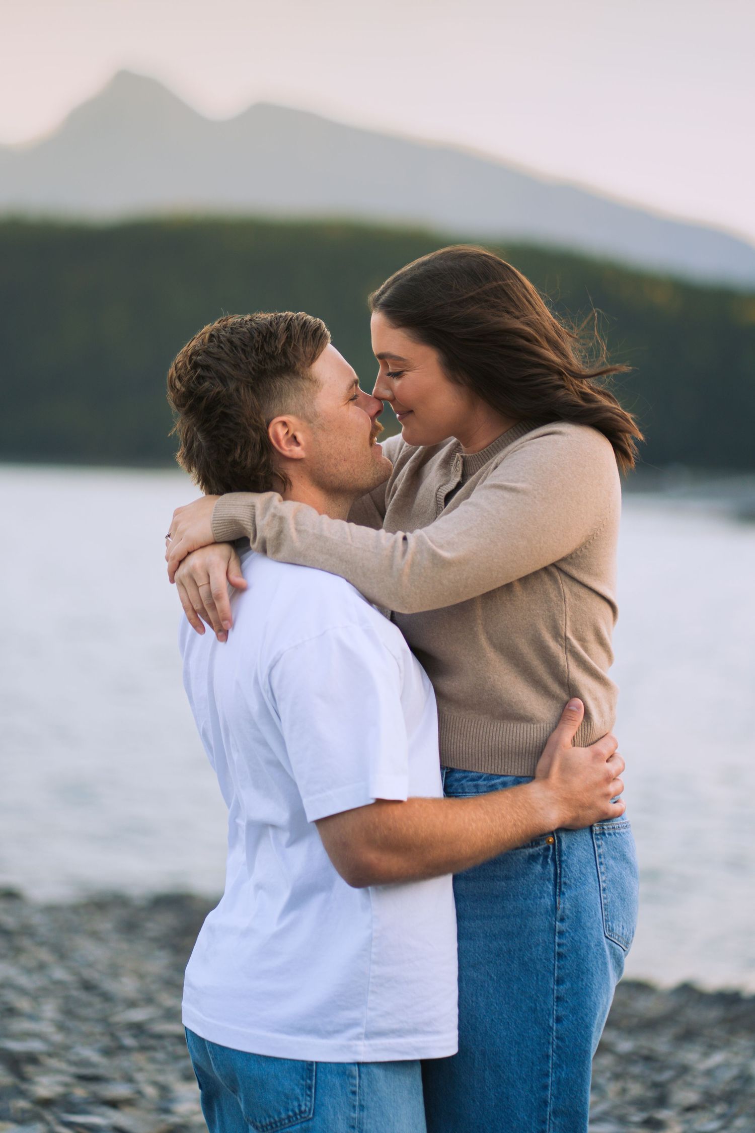 Couple hugging and laughing on the rocky shore at Lake Minnewanka in Banff, with water and forested mountains in the background.