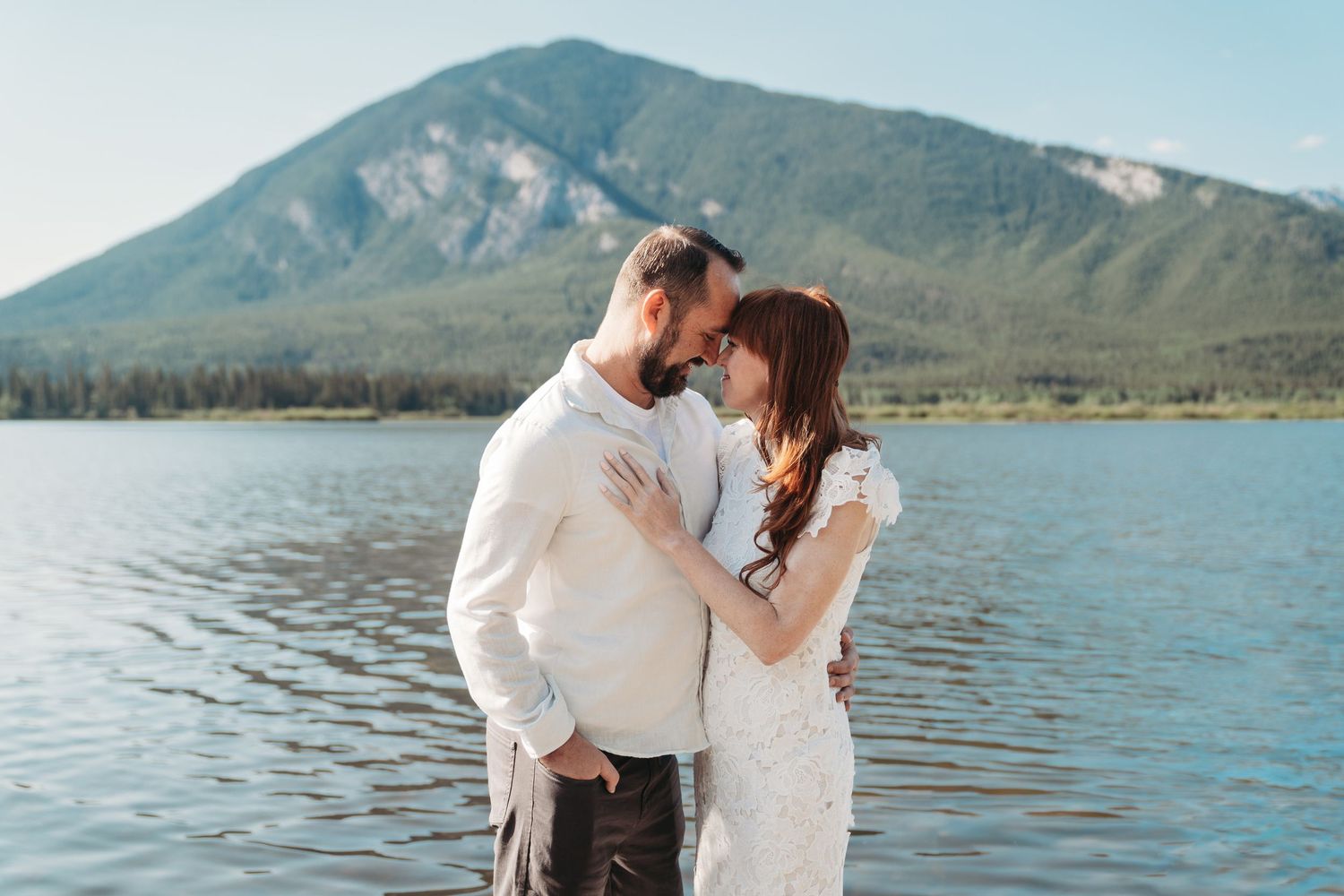 Bride and groom touch foreheads by a calm mountain lake in Banff, sharing an intimate moment with forested peaks in the background.