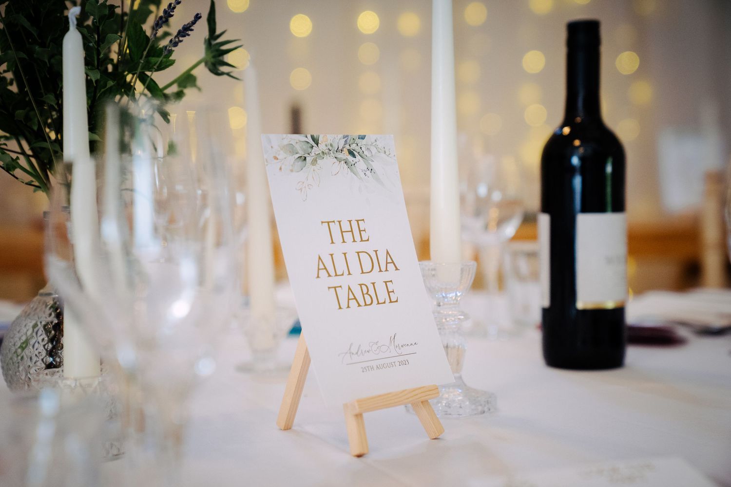 Wedding reception table decor featuring a named table card on an easel with wine, glassware and candles in soft ambient lighting.