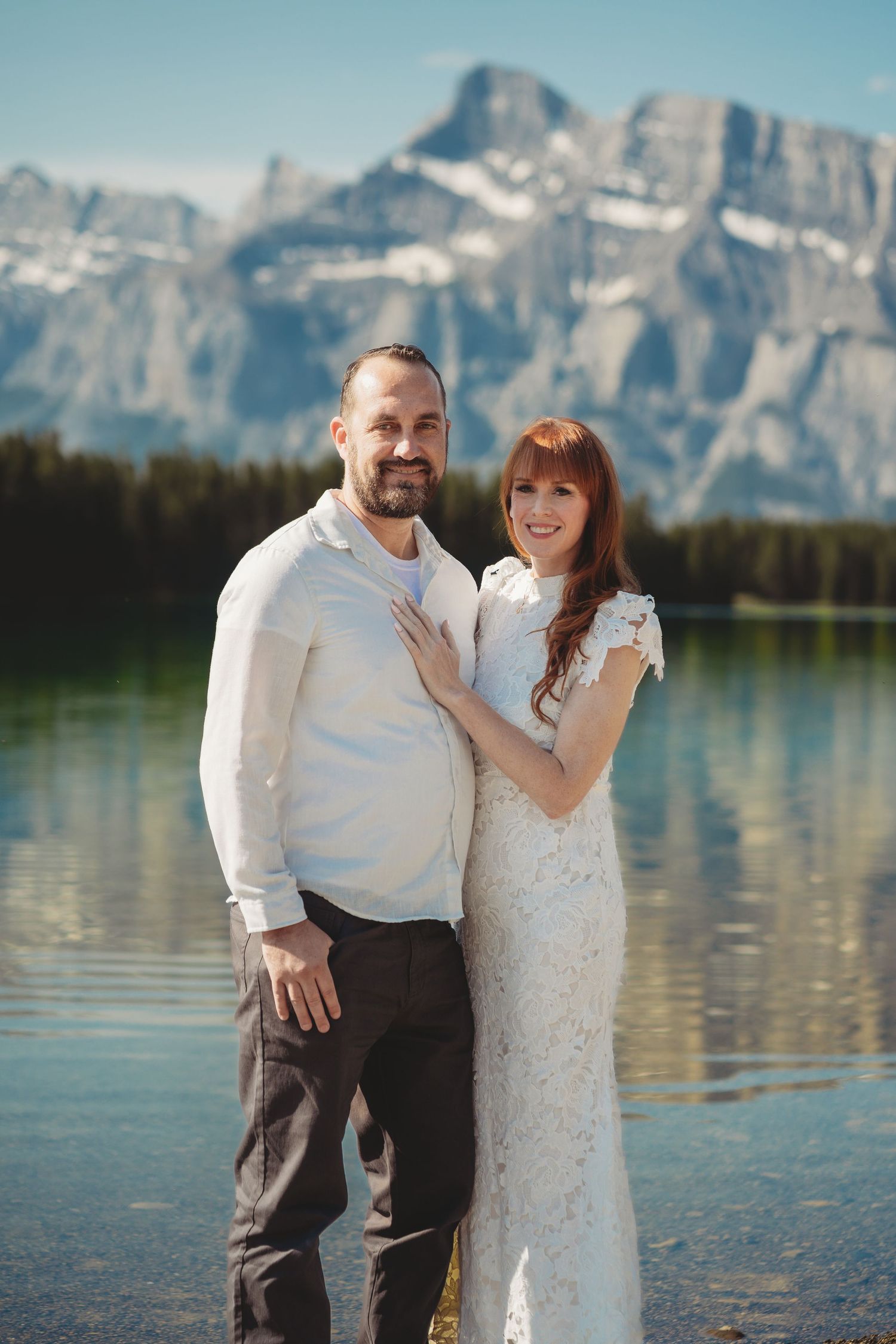Bride and groom stand smiling by a calm mountain lake in Banff, surrounded by alpine peaks, evergreen trees, and clear blue summer skies.