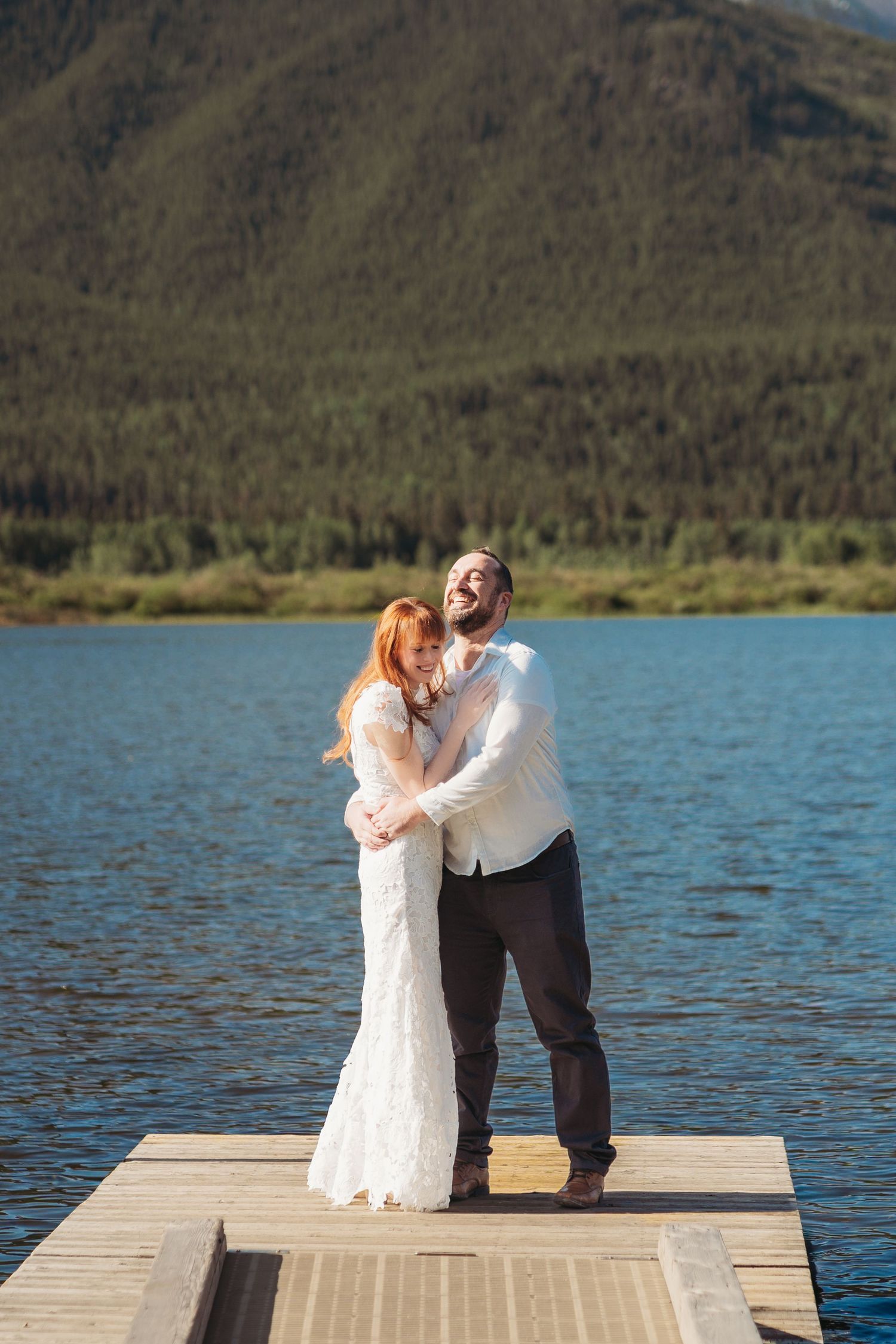Bride and groom laugh together on a dock overlooking a calm mountain lake in Banff, sharing a joyful moment in the sun with forested peaks.