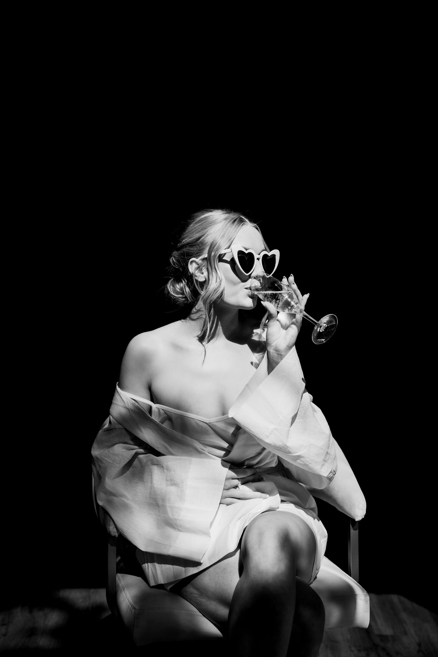 Bride in heart-shaped sunglasses sipping champagne in dramatic black and white lighting, a stylish and playful wedding morning moment.