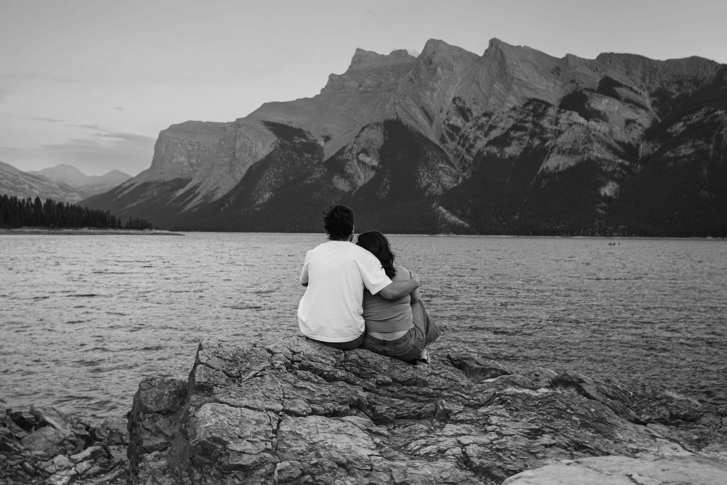 Couple sitting together on rocks facing Lake Minnewanka in Banff, captured in black and white with dramatic mountain peaks across the water.