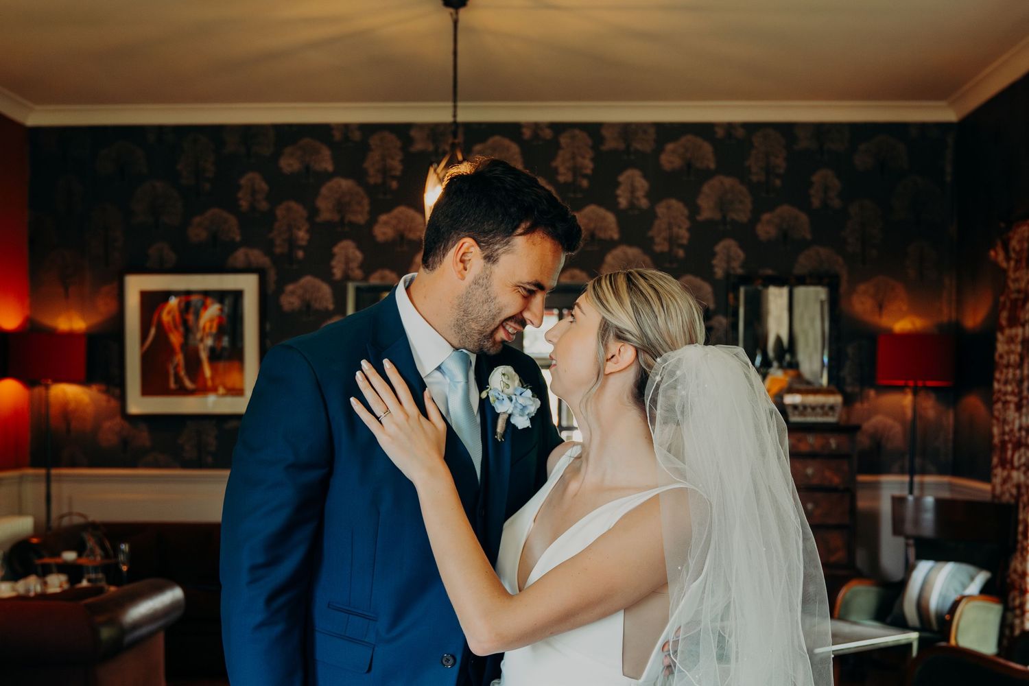 Bride and groom share a joyful, intimate moment indoors, smiling close together in warm light on their wedding day.