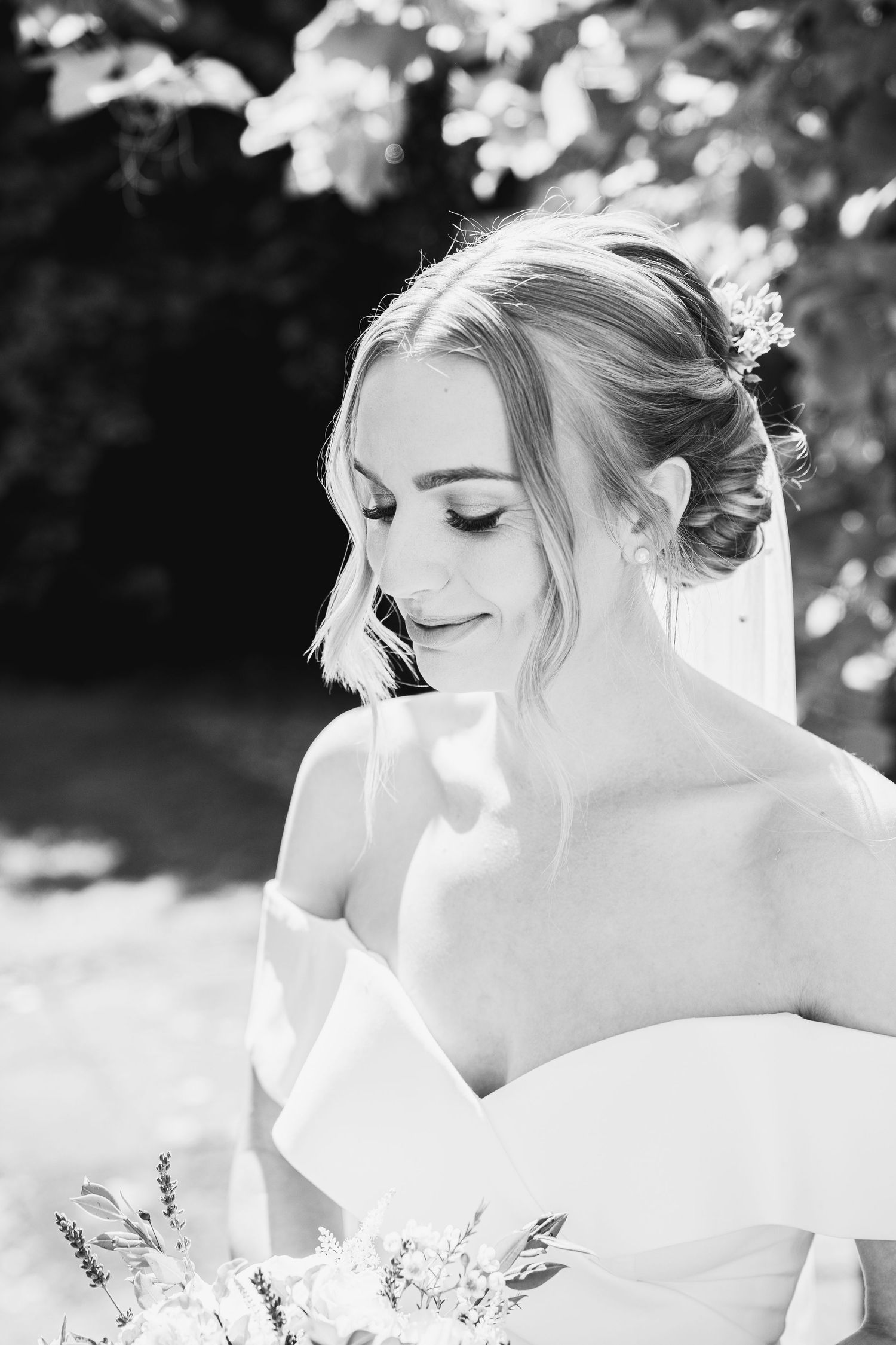 The bride wears a modern off-the-shoulder wedding gown a, photographed outdoors in natural daylight with a soft, gentle smile.