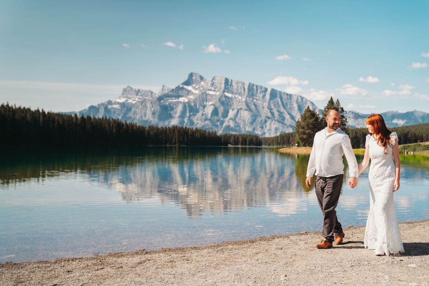 Bride and groom walk hand in hand along a mountain lake in Banff, with clear reflections and stunning rocky peaks in the background.