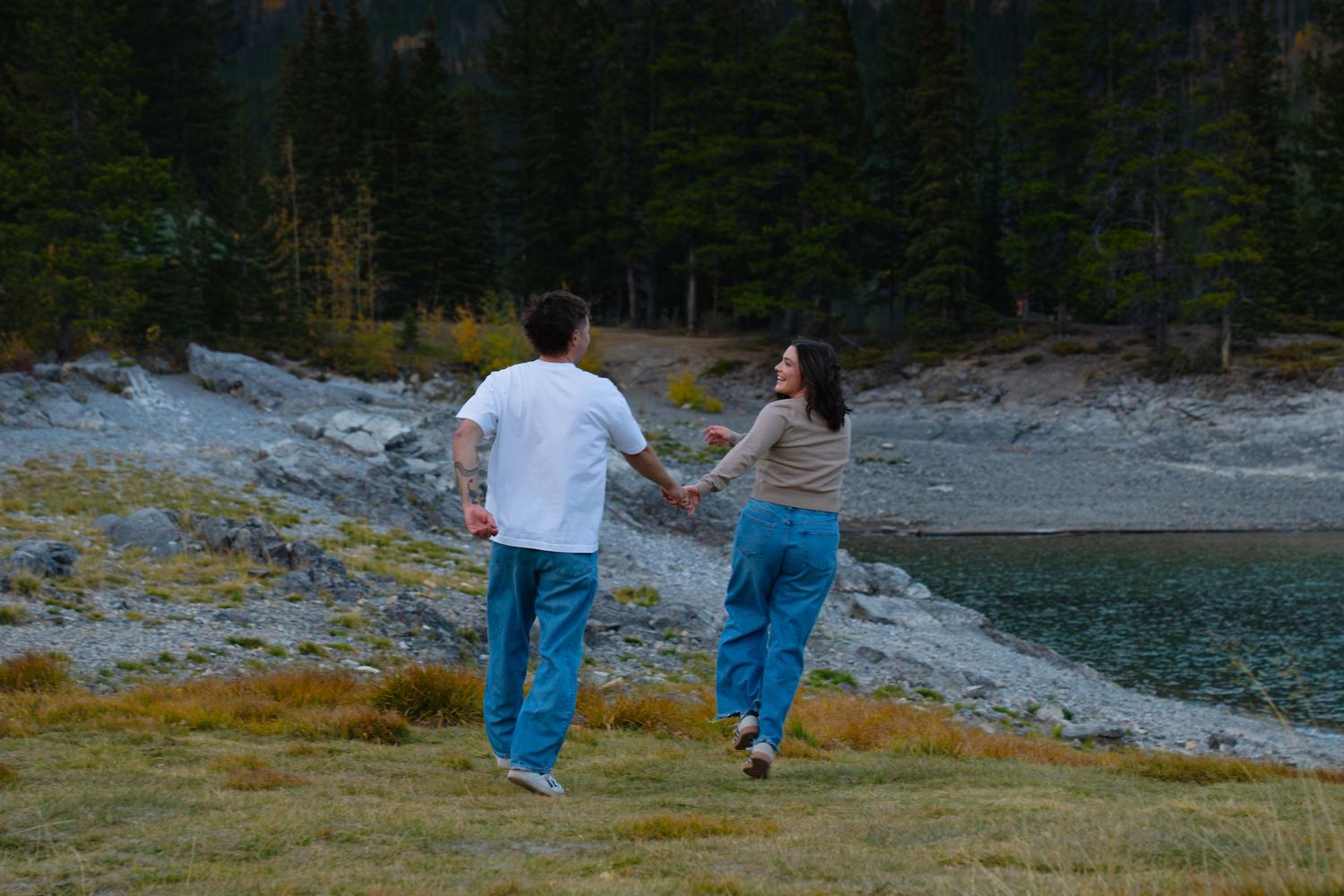 Couple holding hands, running along the shoreline at Lake Minnewanka in Banff, surrounded by forest and rocky landscape during an engagement