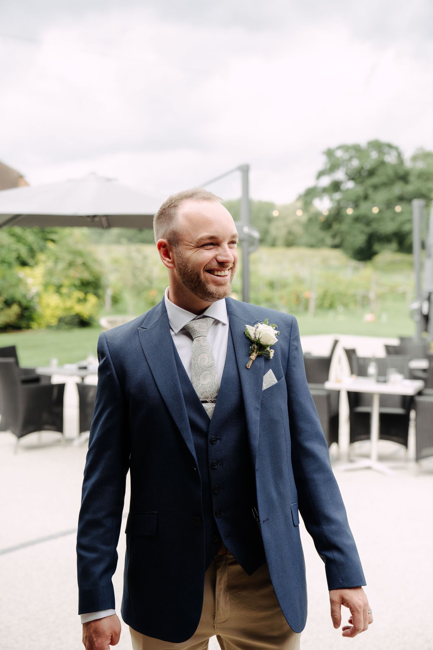 Groom smiling outdoors in a navy suit with boutonniere, captured candidly before the wedding meal. patio seating in the background