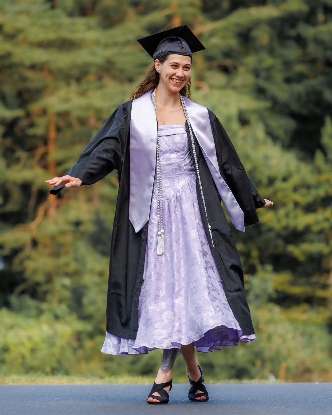 A young woman in black graduation cap and gown over a lavender dress smiles outdoors with trees in background.