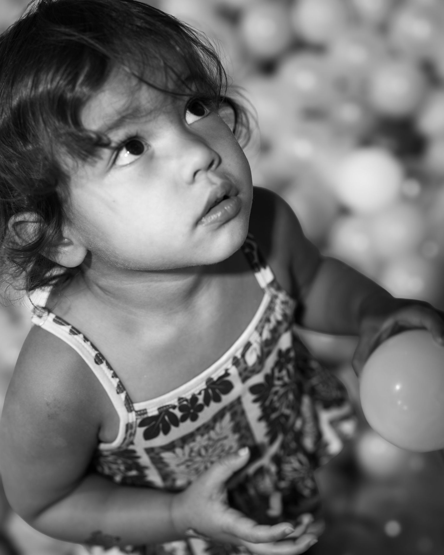 Black and white portrait of a curious toddler girl in a floral dress looking up, surrounded by colorful play balls.