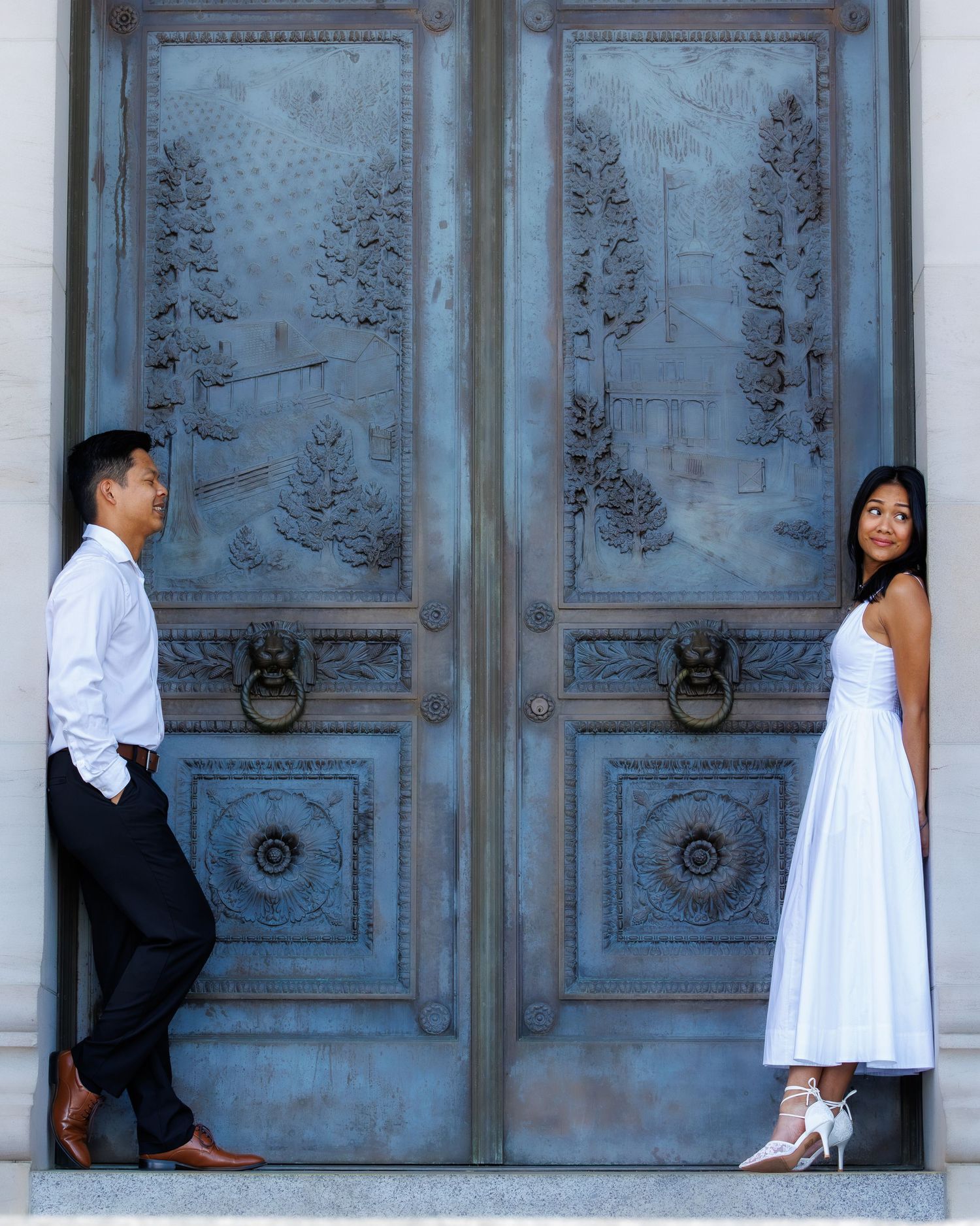 Couple in white outfits posing on either side of ornate blue bronze doors with nature relief carvings.
