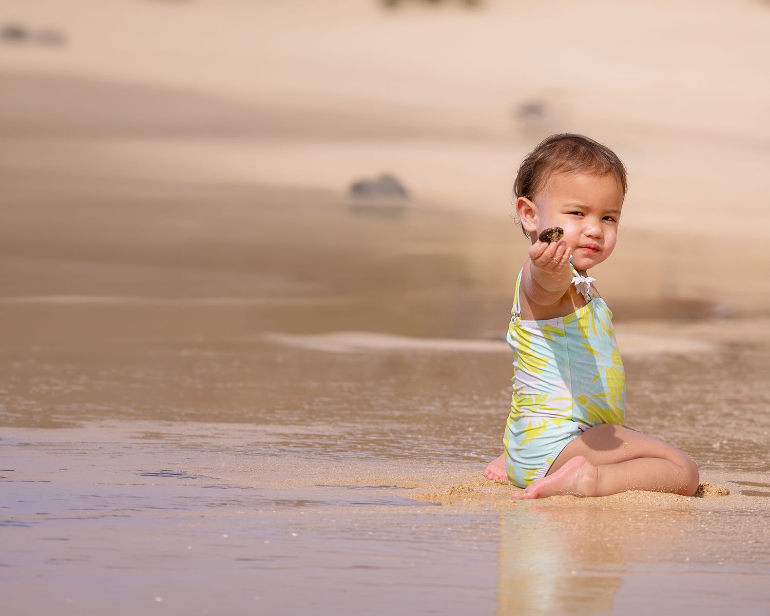 A toddler in a colorful swimsuit sits on a sandy beach, reaching out while waves gently wash around them.