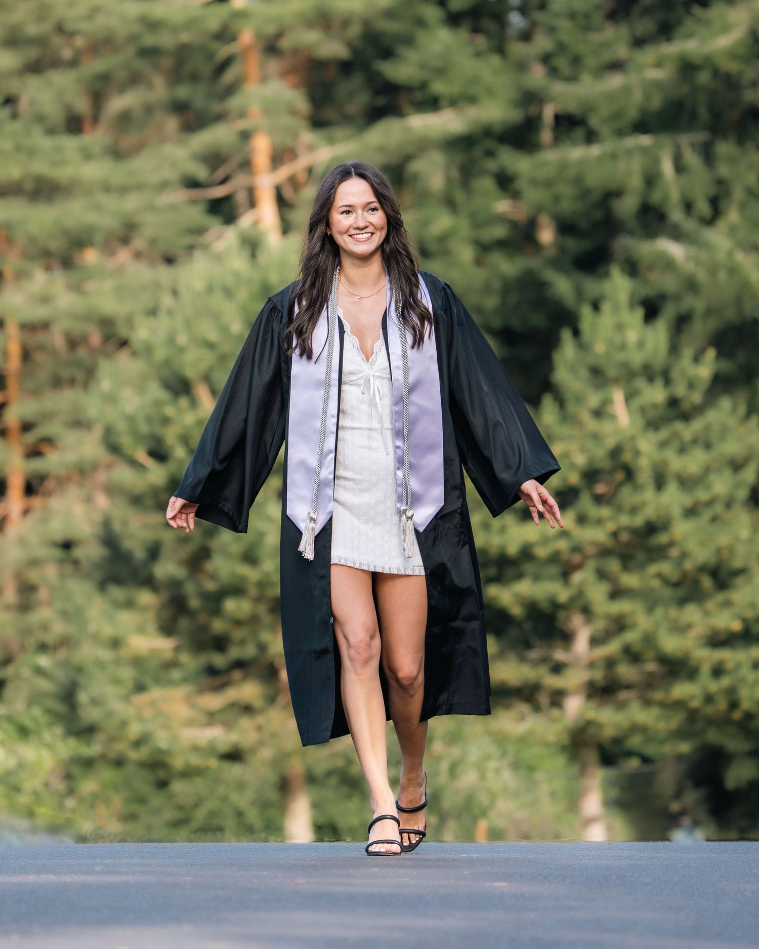 Smiling graduate walks in black cap and gown over white dress, with green trees in background.