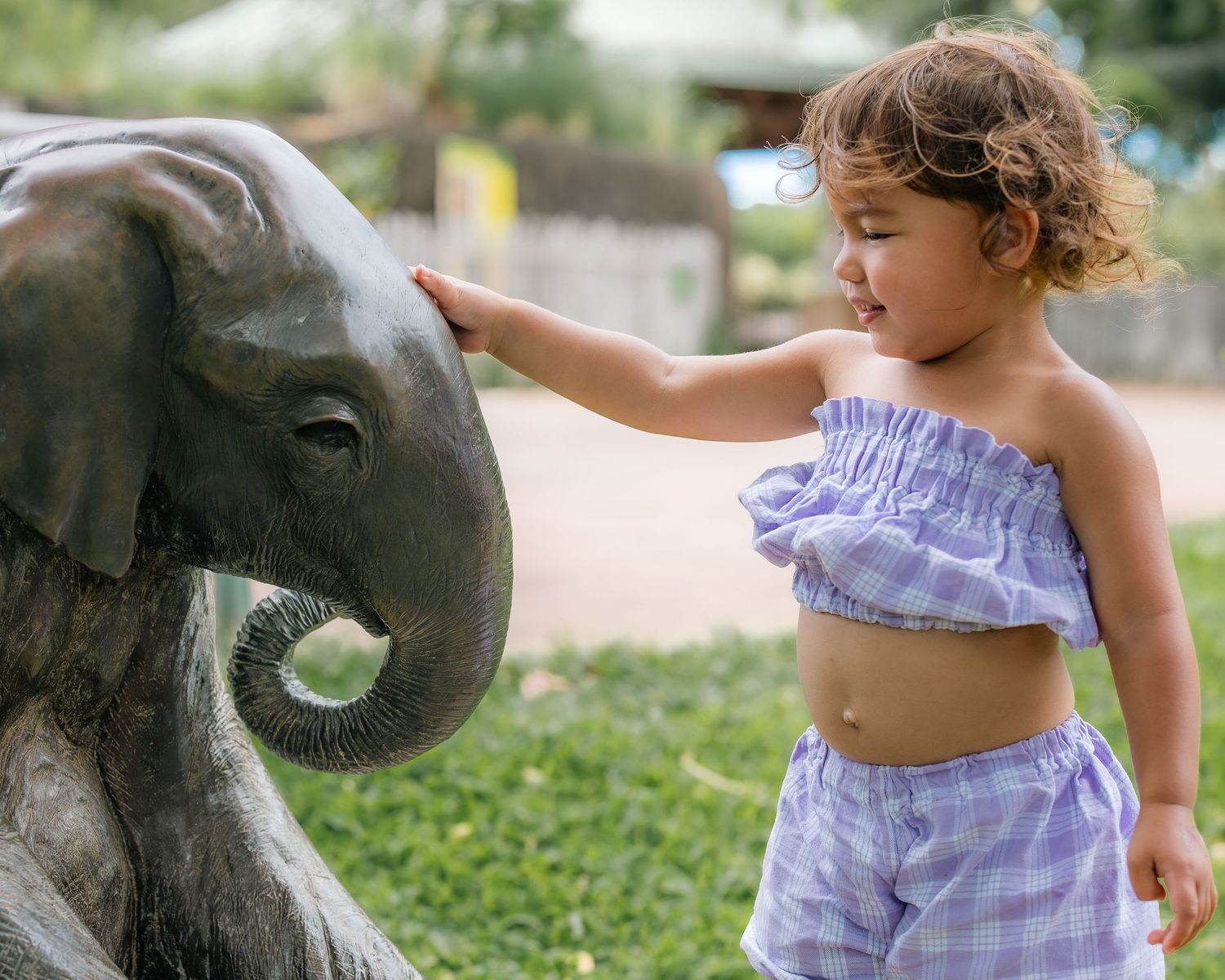 A young toddler girl in a plaid outfit touching a bronze elephant statue at a zoo or park.
