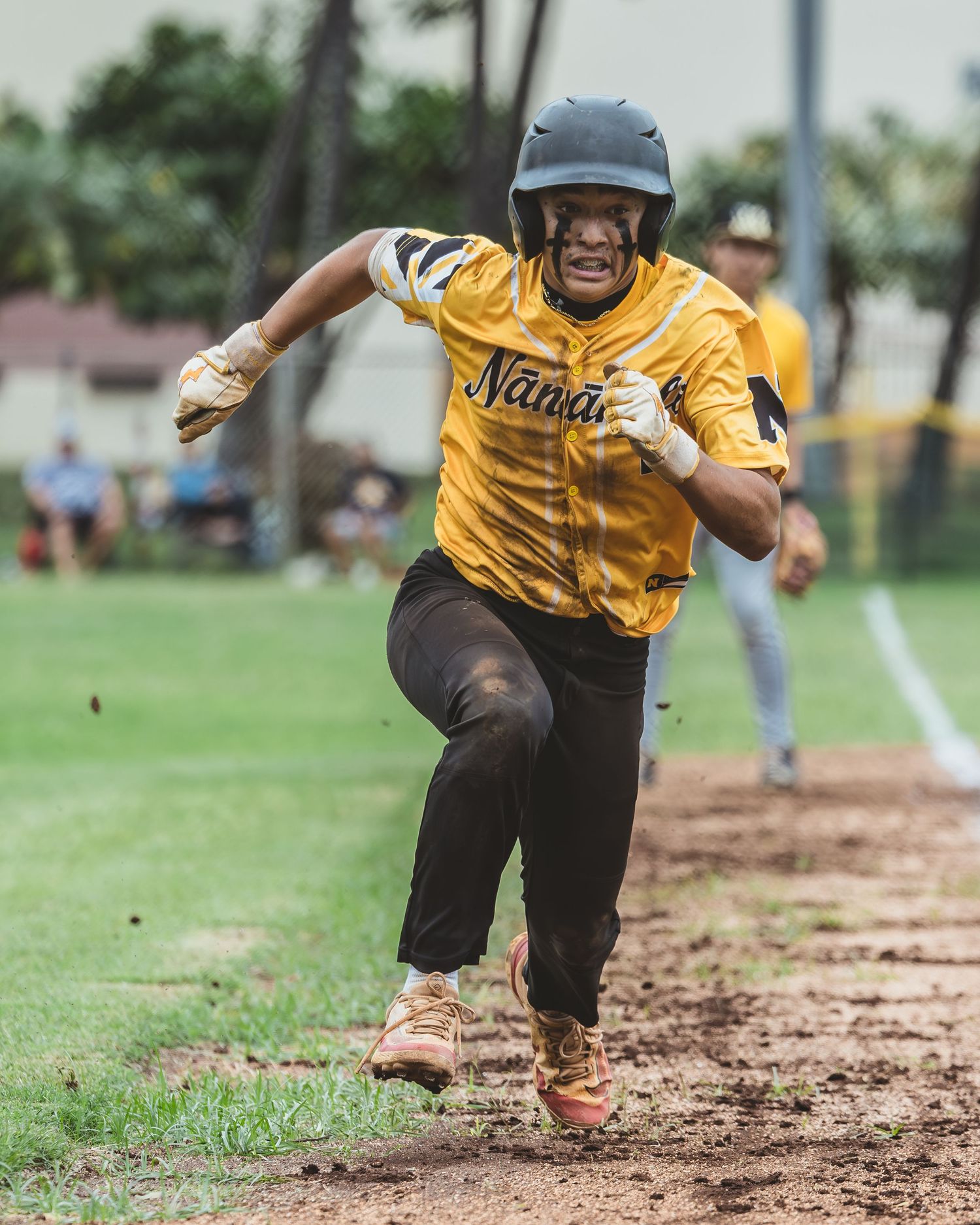 Baseball player in yellow Nanai jersey sprinting on dirt baseline wearing helmet during youth league game.