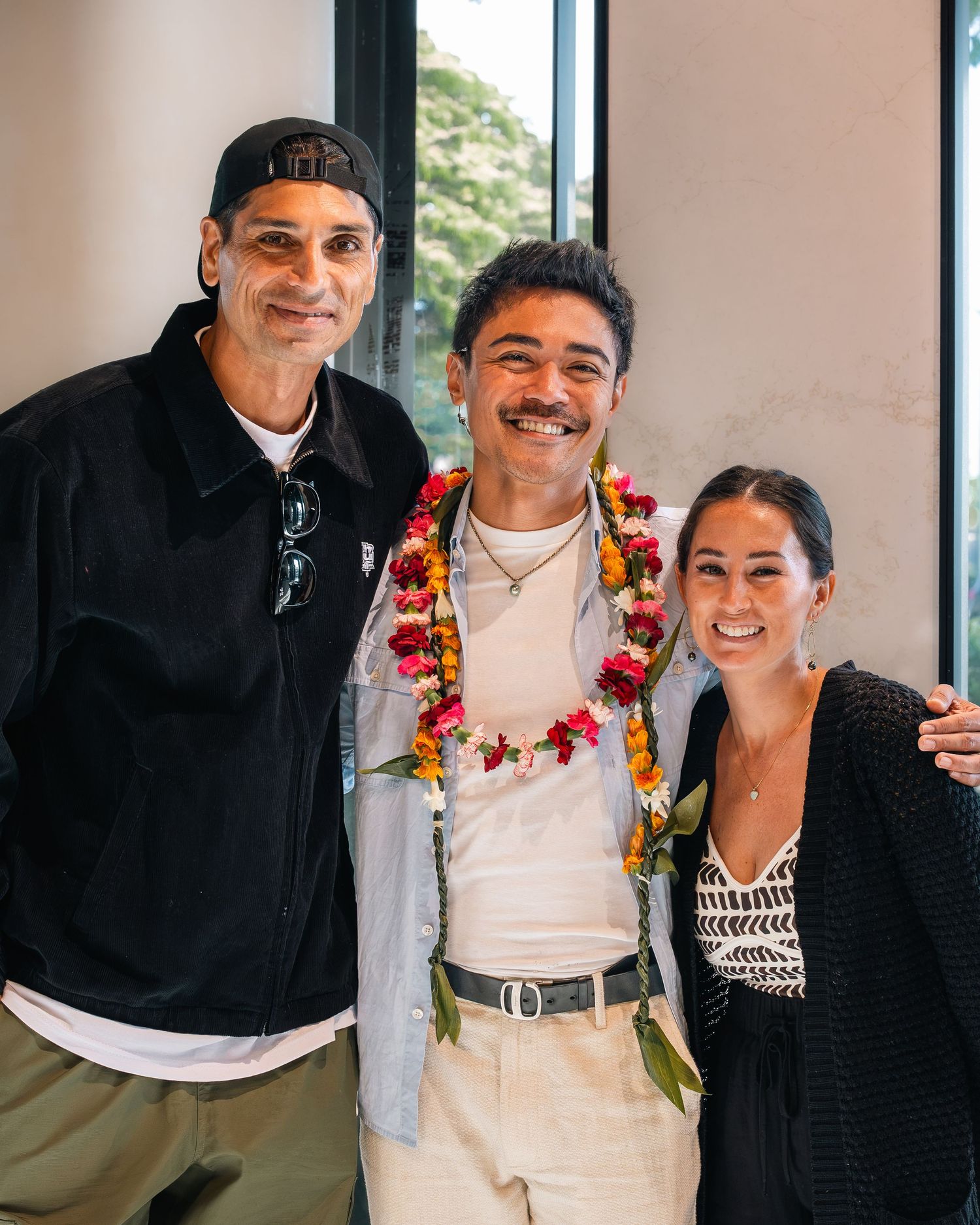 Three people smiling together, middle person wearing a colorful floral lei, posing indoors near a window.