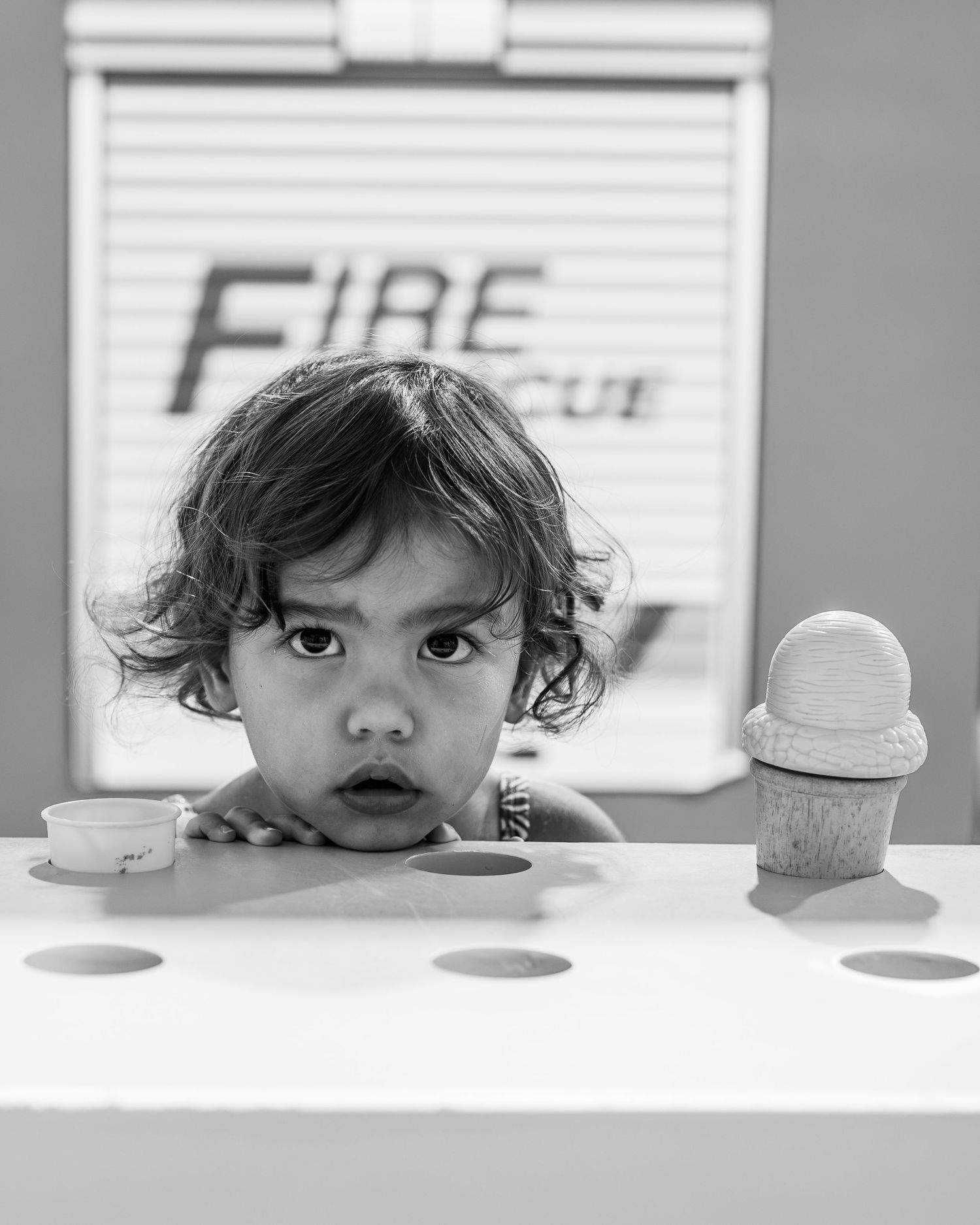 Black and white photo of a curious toddler peering over a white table with toy ice cream and cups, Fire Rescue sign behind.