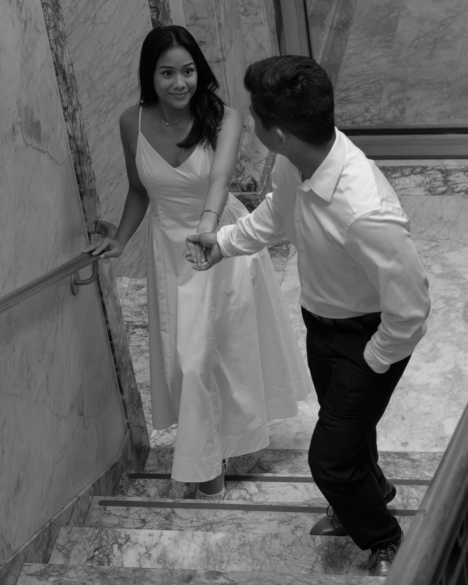 Black and white photo of a couple on marble stairs, woman in white dress smiling at man in white shirt.