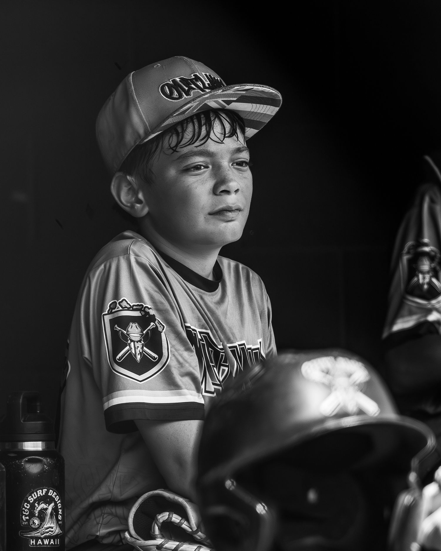 Black and white photo of a young boy in baseball uniform and cap sitting in dugout with helmets nearby.