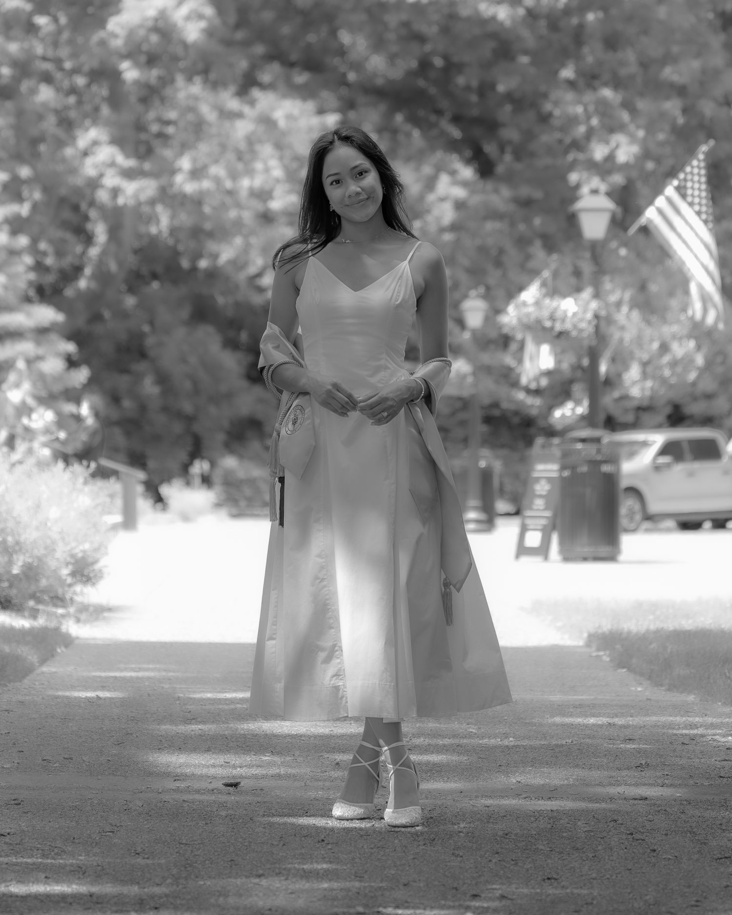 Young woman in midi dress and heels poses on a tree-lined path with an American flag in the background.