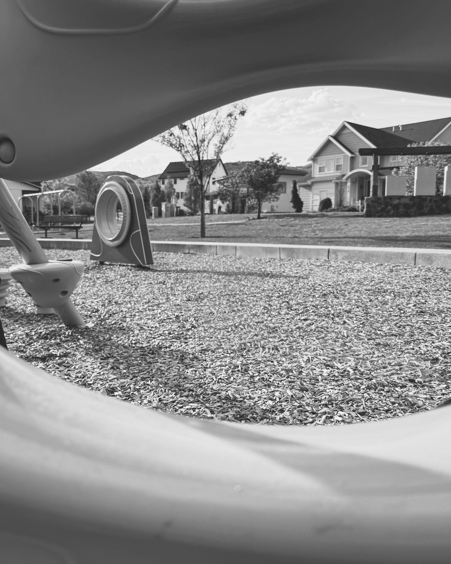 Black and white playground slide view overlooking a suburban neighborhood with houses, trees, and wood chip ground cover.