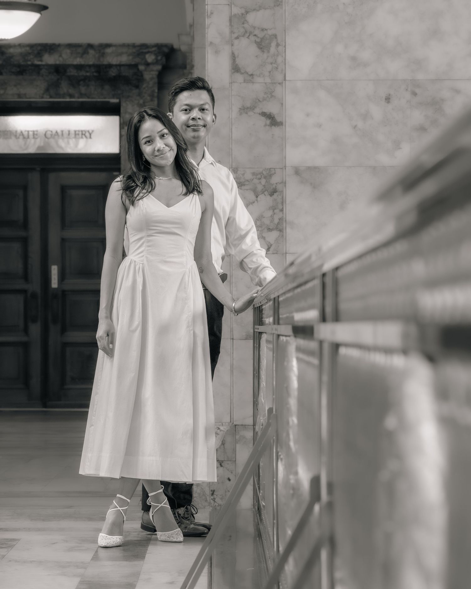 Black and white photo of a couple in formal wear holding hands inside an elegant gallery building.