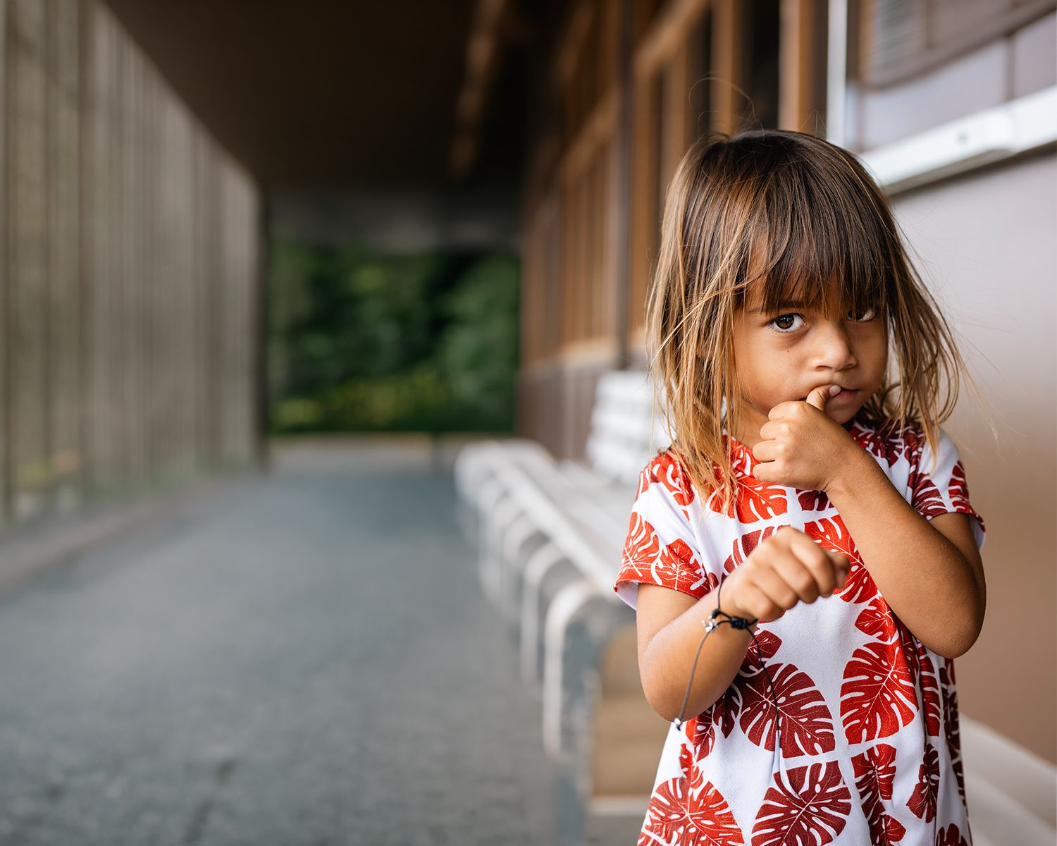 Young girl in red floral dress standing in a hallway corridor, looking thoughtful with hands raised.