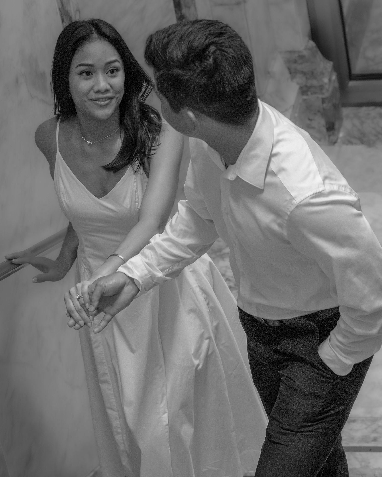 Black and white photo of a couple holding hands on stairs, woman in white dress smiling at man in white shirt.