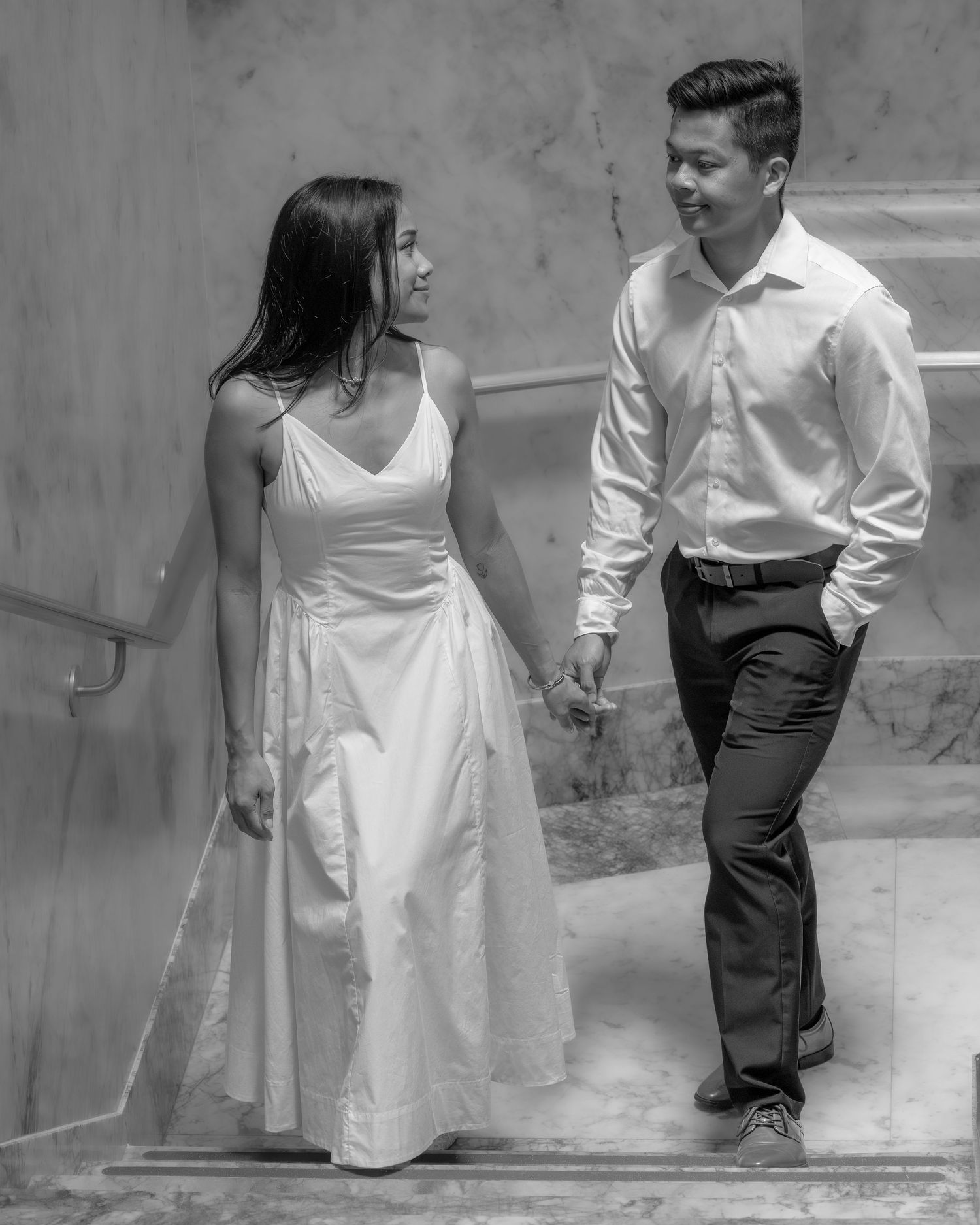 Black and white photo of a couple holding hands on stairs, woman in white dress, man in white shirt and dark pants.