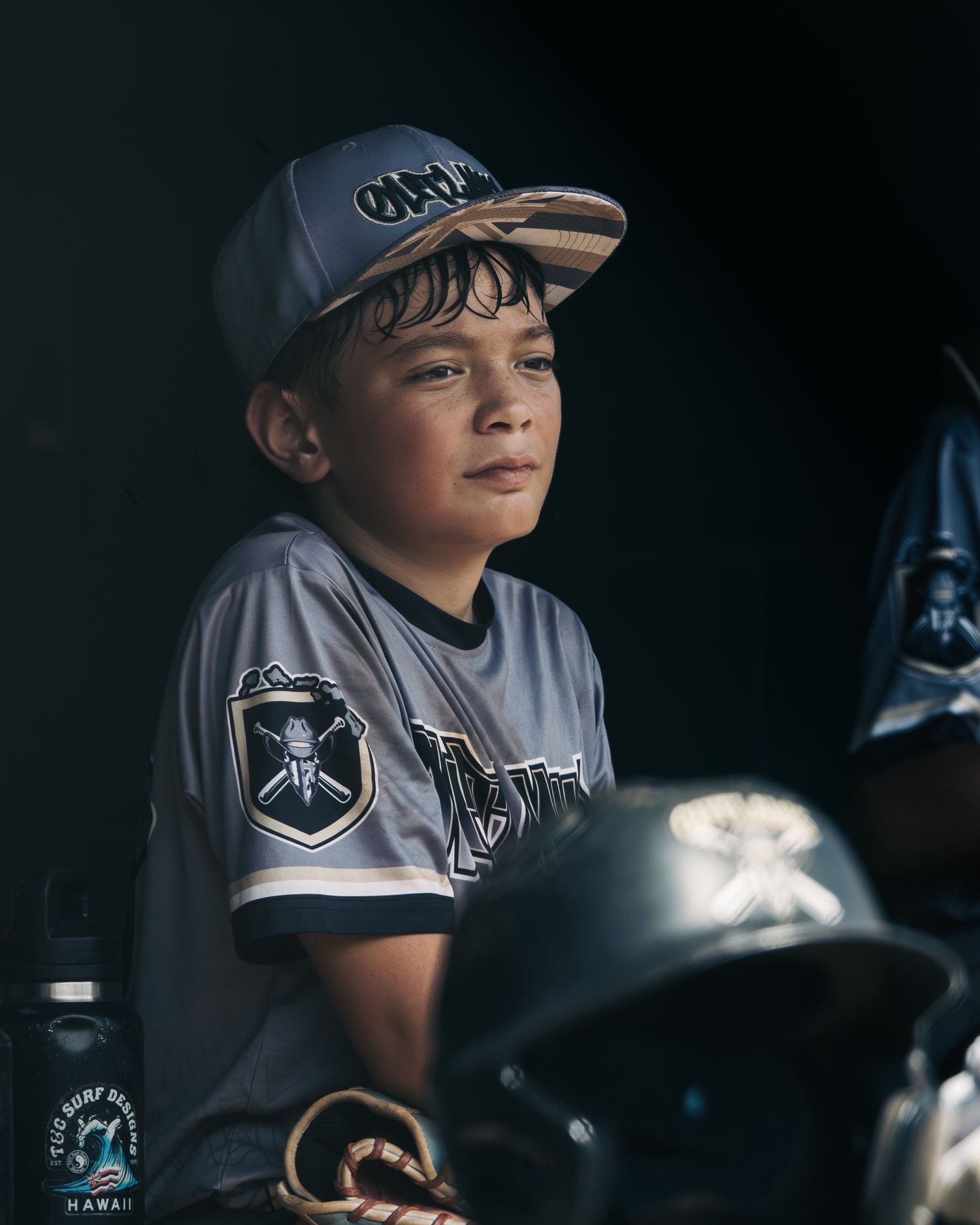 Young boy in baseball uniform and cap sits in dugout, looking focused during a little league game in Hawaii.