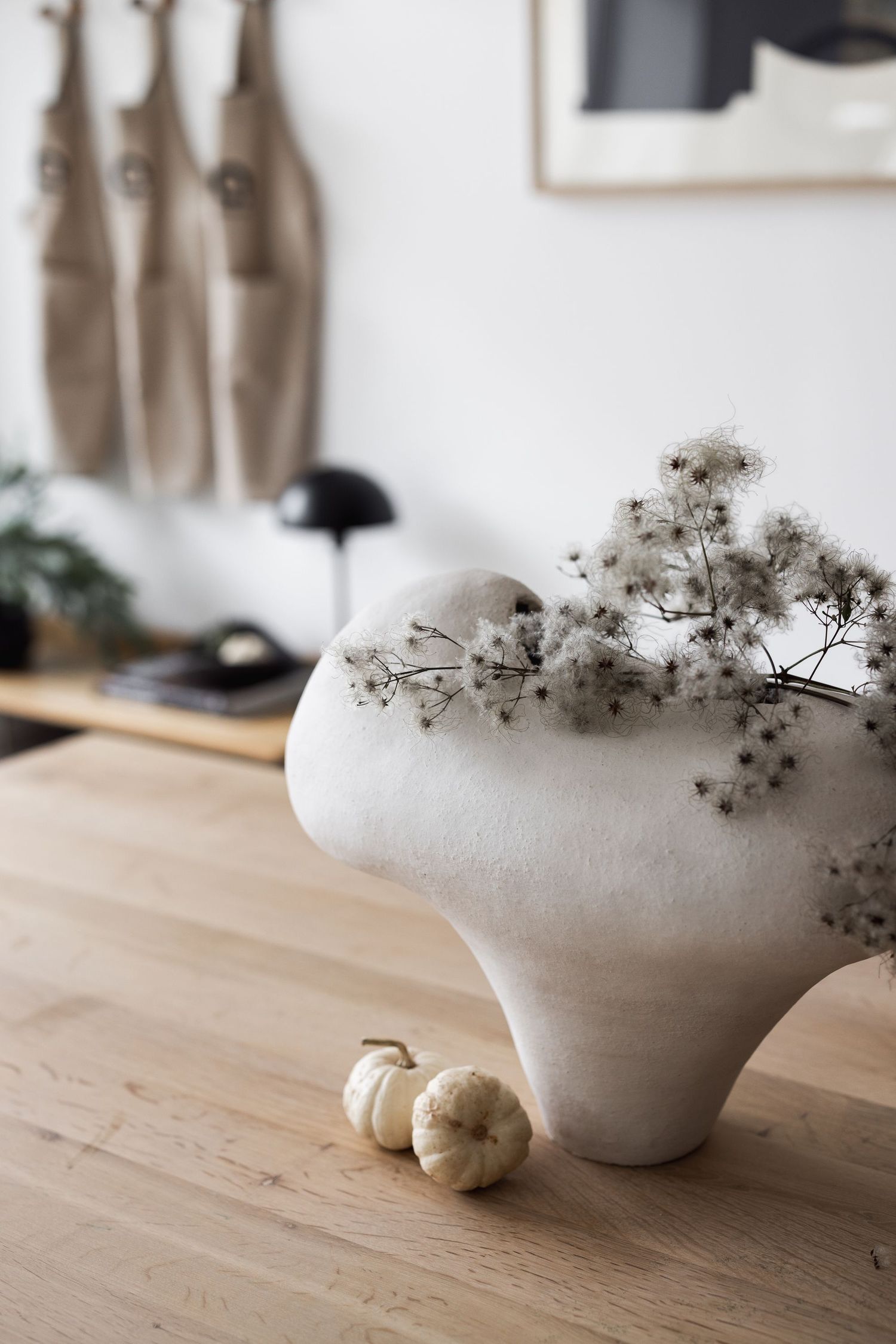 White sculptural vase with dried flowers and mini pumpkins on a wooden table in a minimalist Scandinavian interior.
