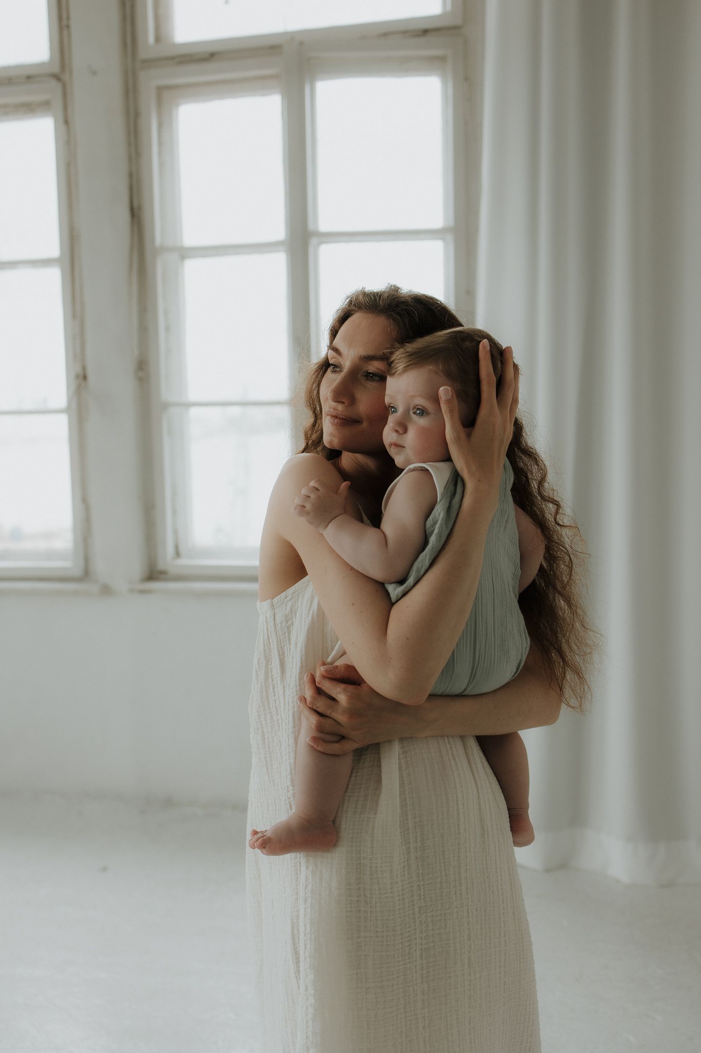Mother with curly hair in white dress tenderly holding her baby in a bright room.