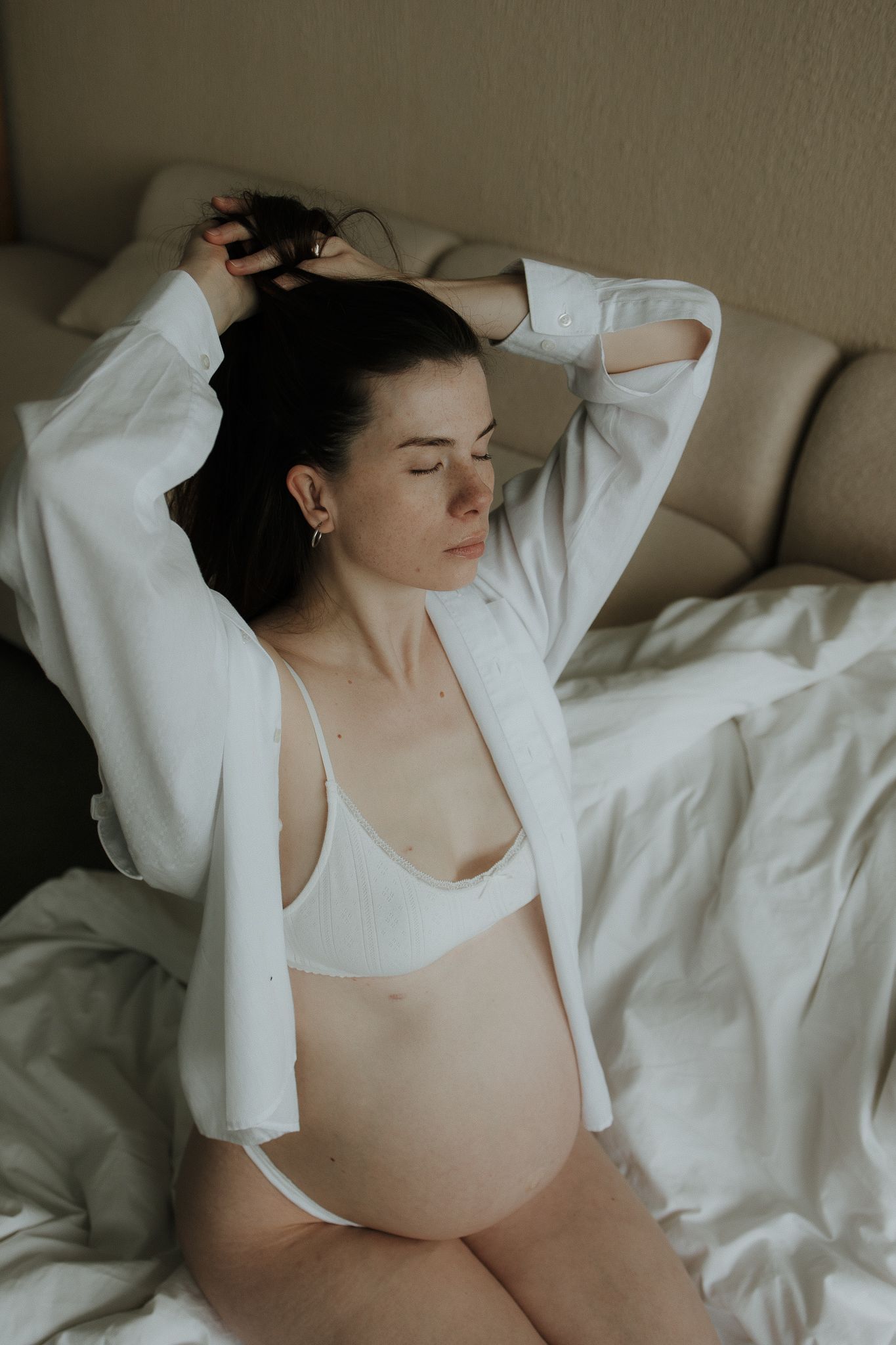 Pregnant woman in white lingerie and open shirt sitting on a bed with eyes closed and hands in hair.