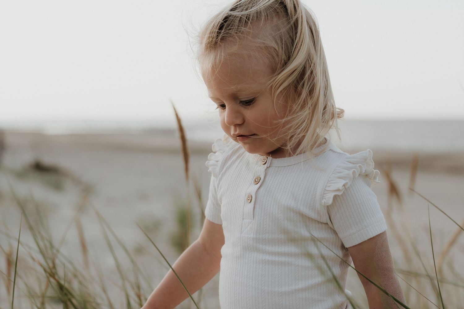 A young blonde girl in a white ruffled top looks down while standing among beach dune grass.