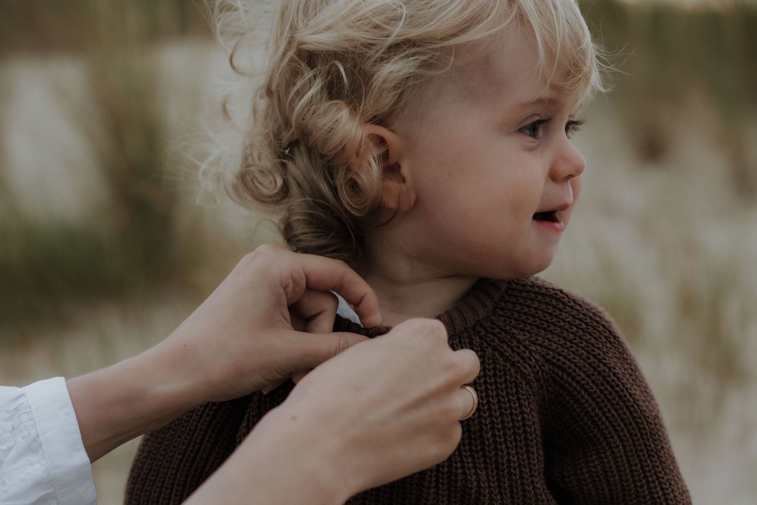 Mother fastening brown knit sweater on curly blonde toddler in sandy outdoor setting.