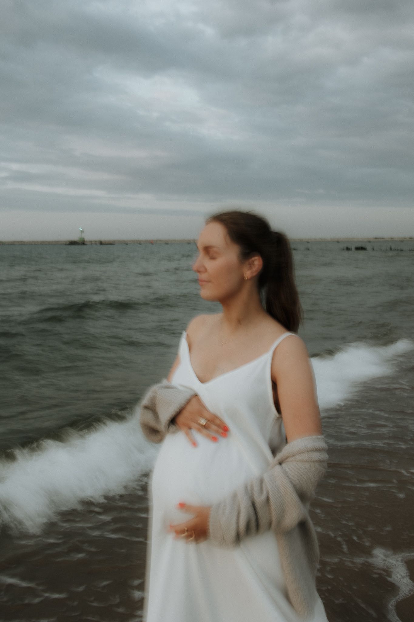 Pregnant woman in white dress and cardigan standing in ocean waves on cloudy beach day.