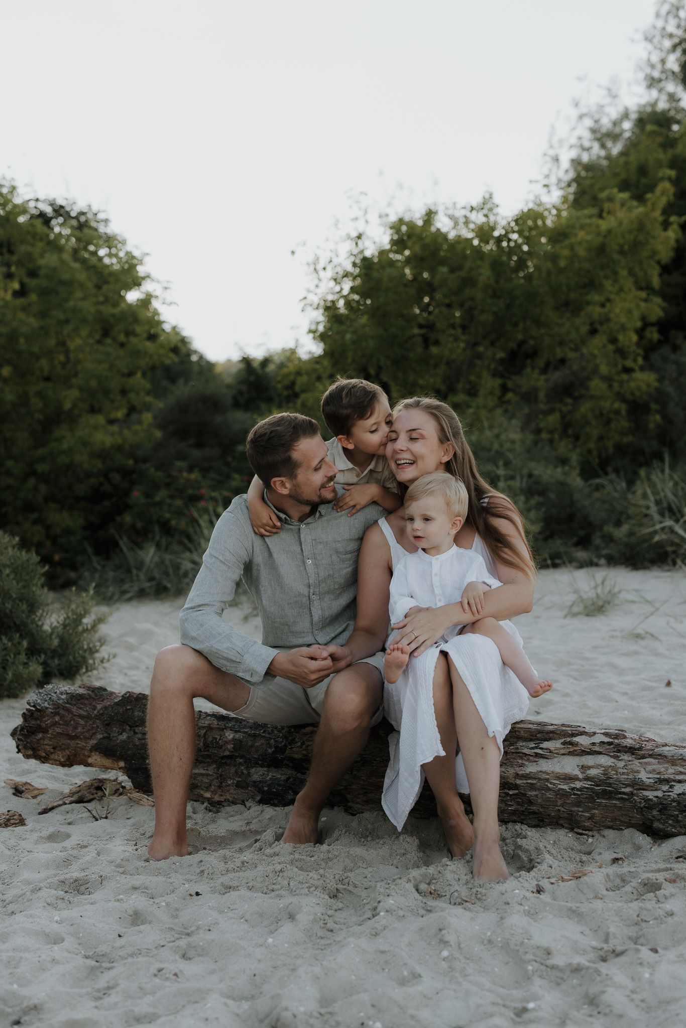 Happy family of four sitting on a driftwood log on a sandy beach surrounded by green trees.