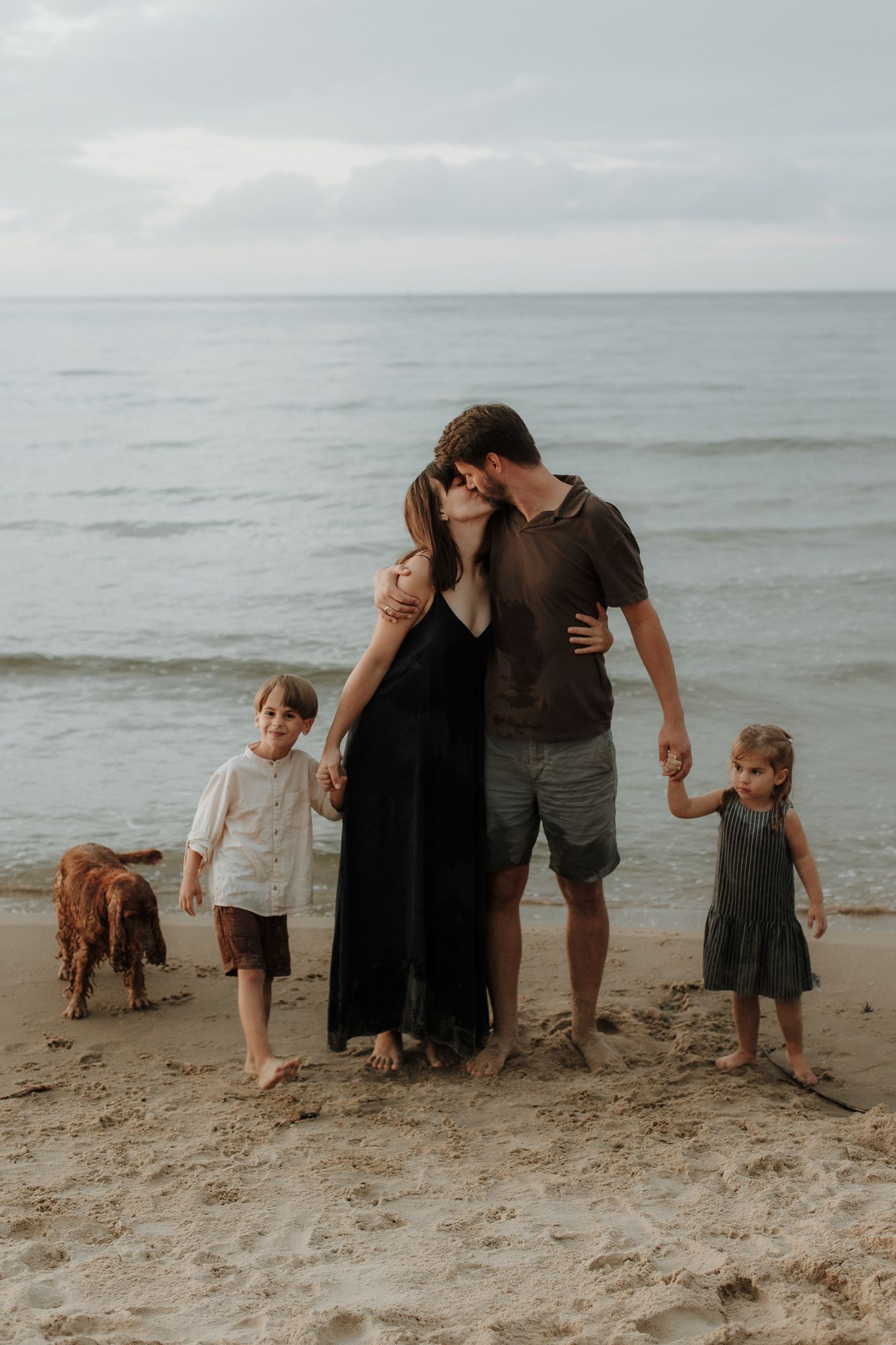 Family of four with a dog sharing a kiss on a cloudy beach by the ocean.