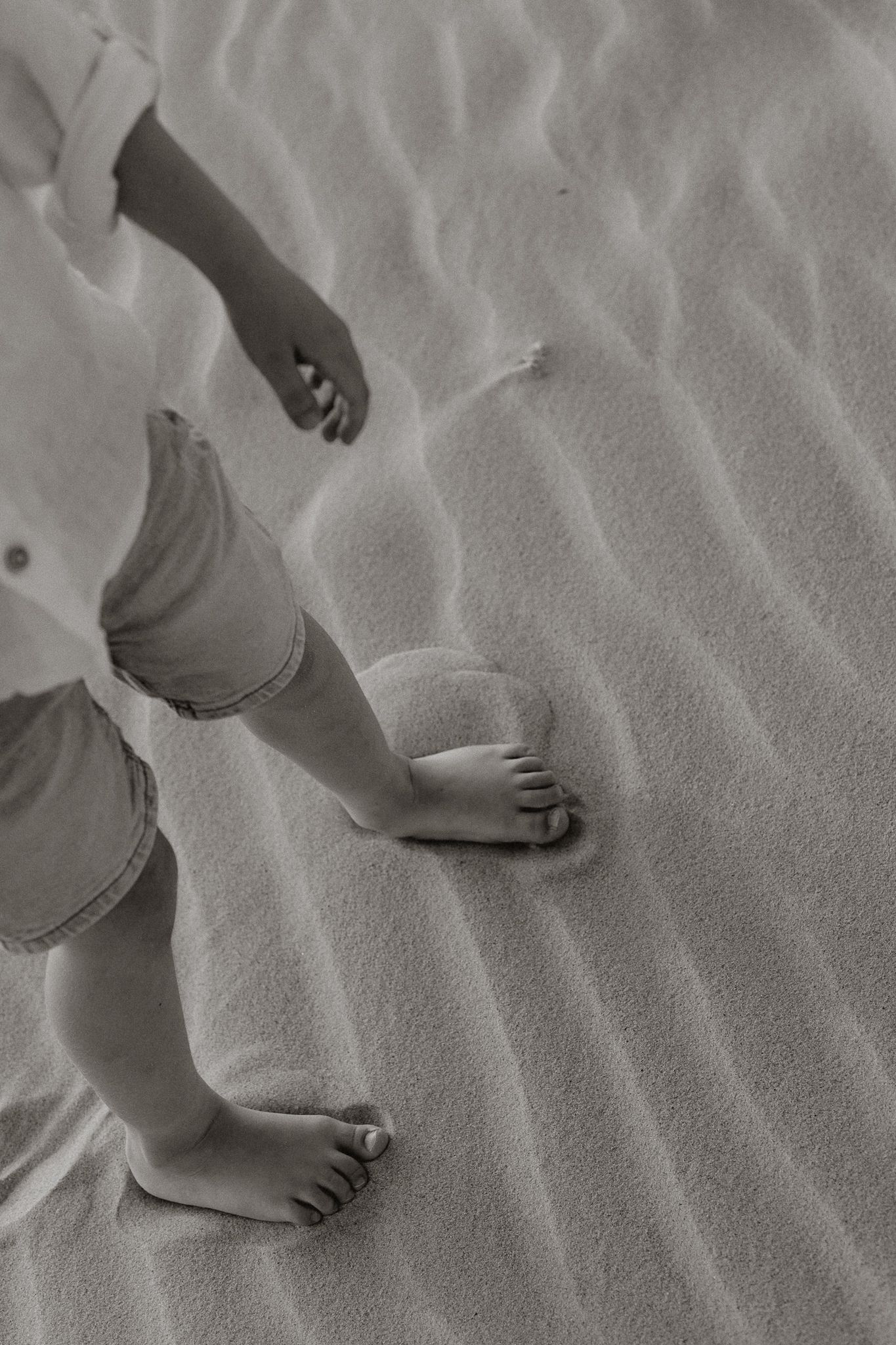 Black and white close-up of a child's bare feet and legs walking on rippled sand.