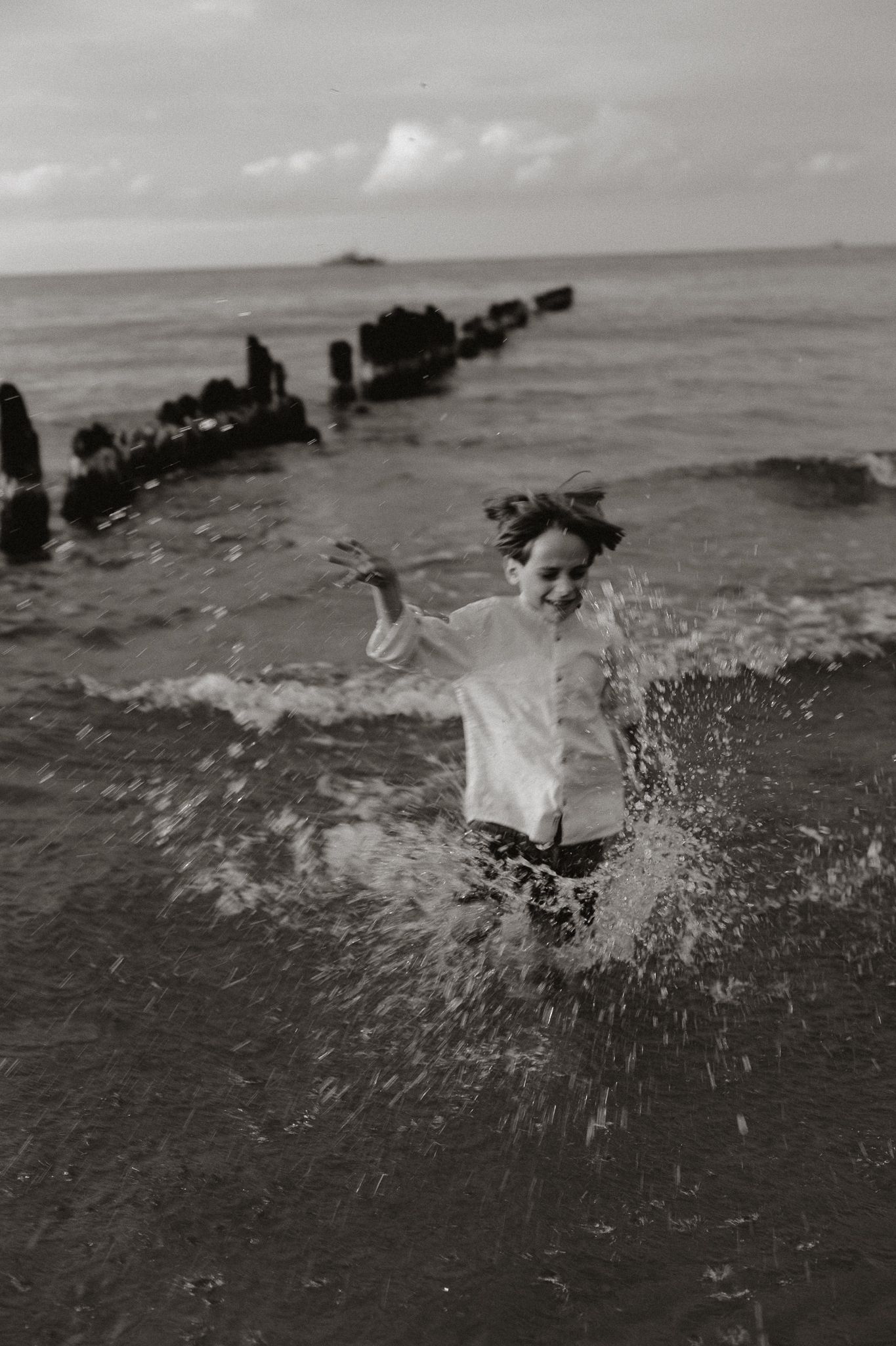 Black and white photo of joyful child splashing through ocean waves on a rocky shore.