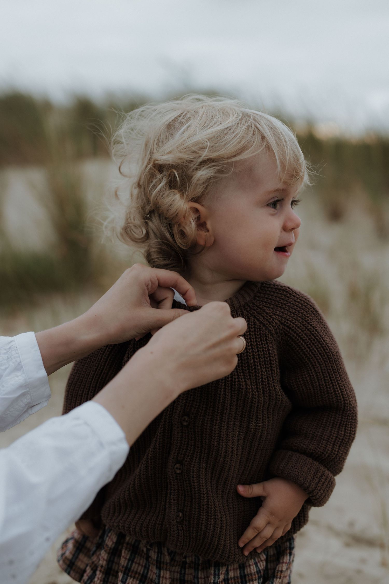 Mother gently buttoning brown knit sweater on curly-haired toddler standing on sandy beach.