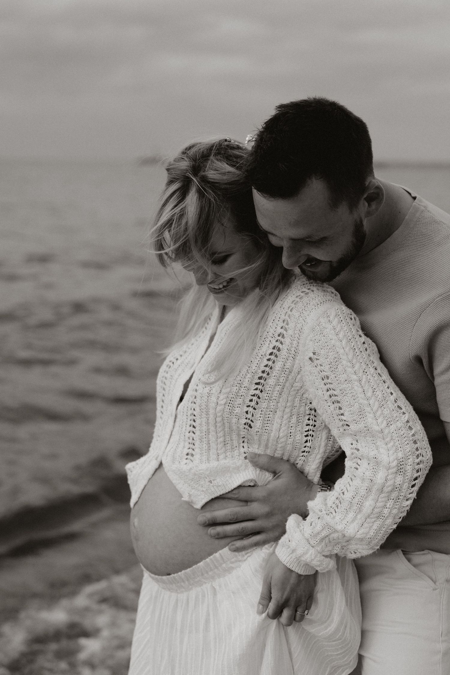 Black and white photo of couple embracing on beach, man hugging pregnant woman from behind.