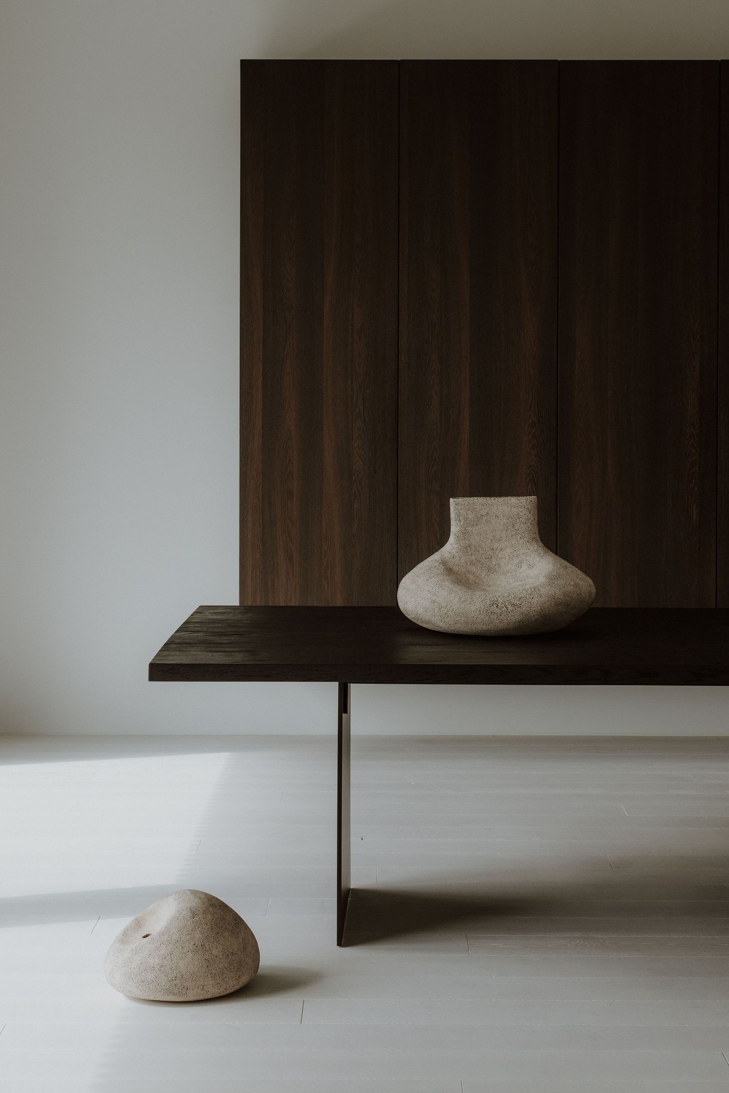 Minimalist interior with dark wood console table, textured ceramic vase, and stone sculpture on white floor.