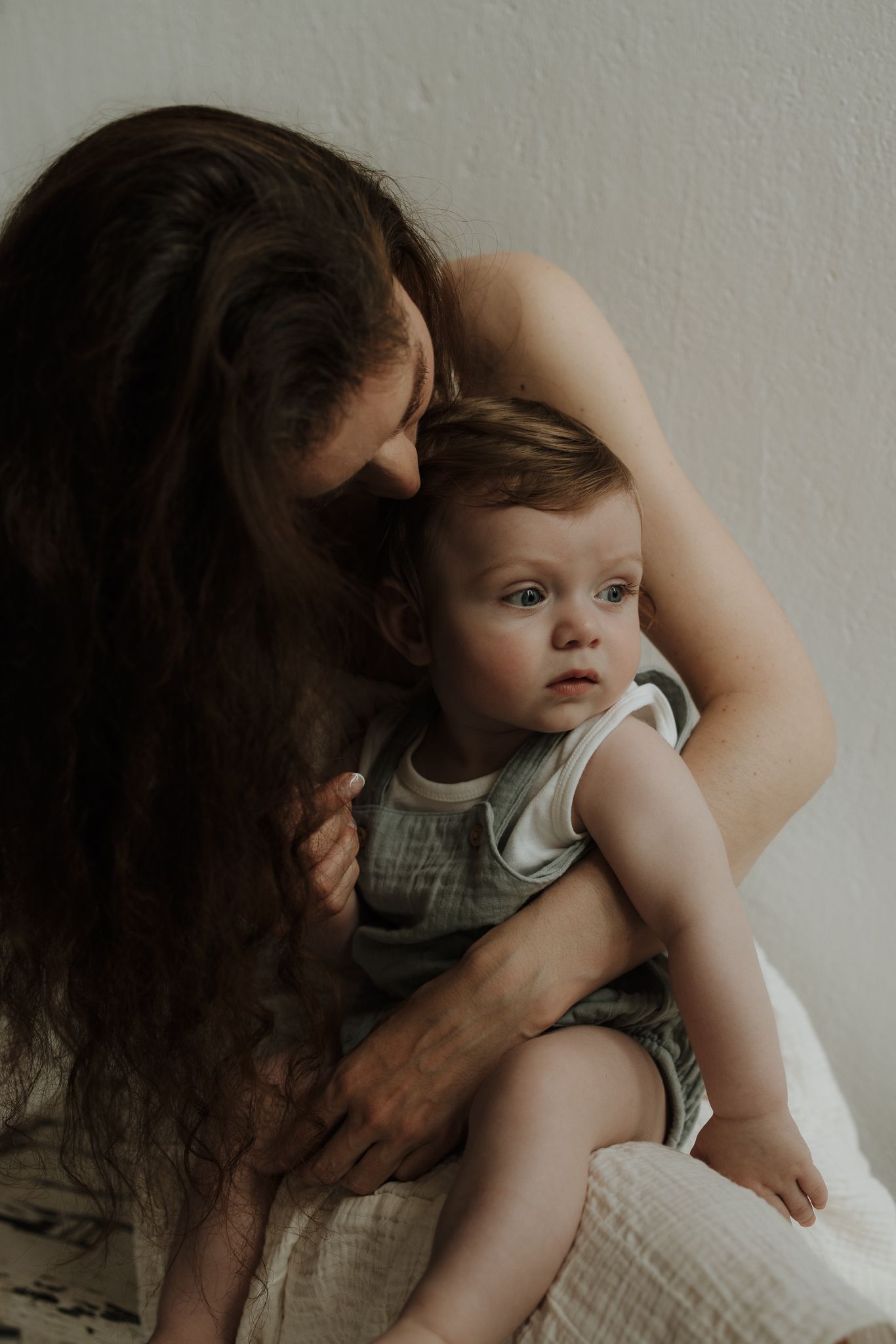 Mother with long curly hair tenderly holding and kissing her baby in a soft white romper.