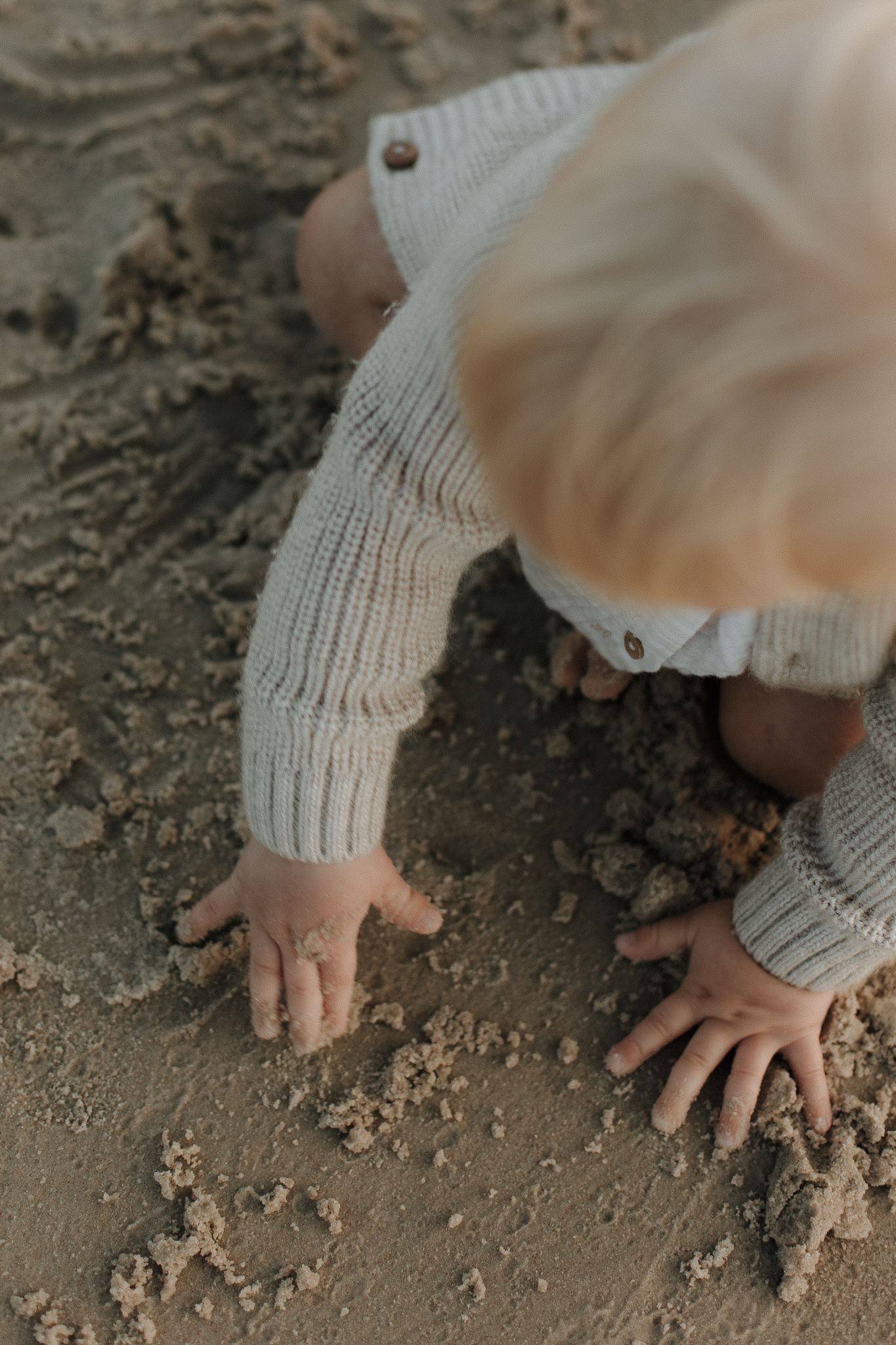 Close-up of a baby in a knit sweater crawling and exploring wet sand at the beach.