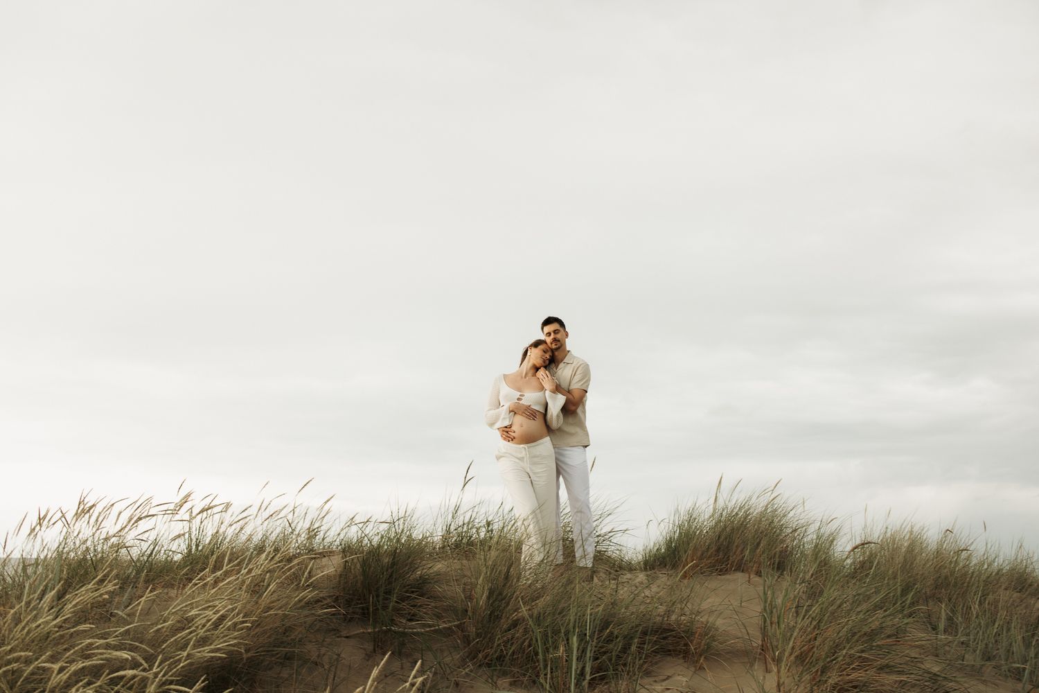Pregnant couple standing together on a sandy dune with beach grass, sharing an intimate embrace in white outfits.